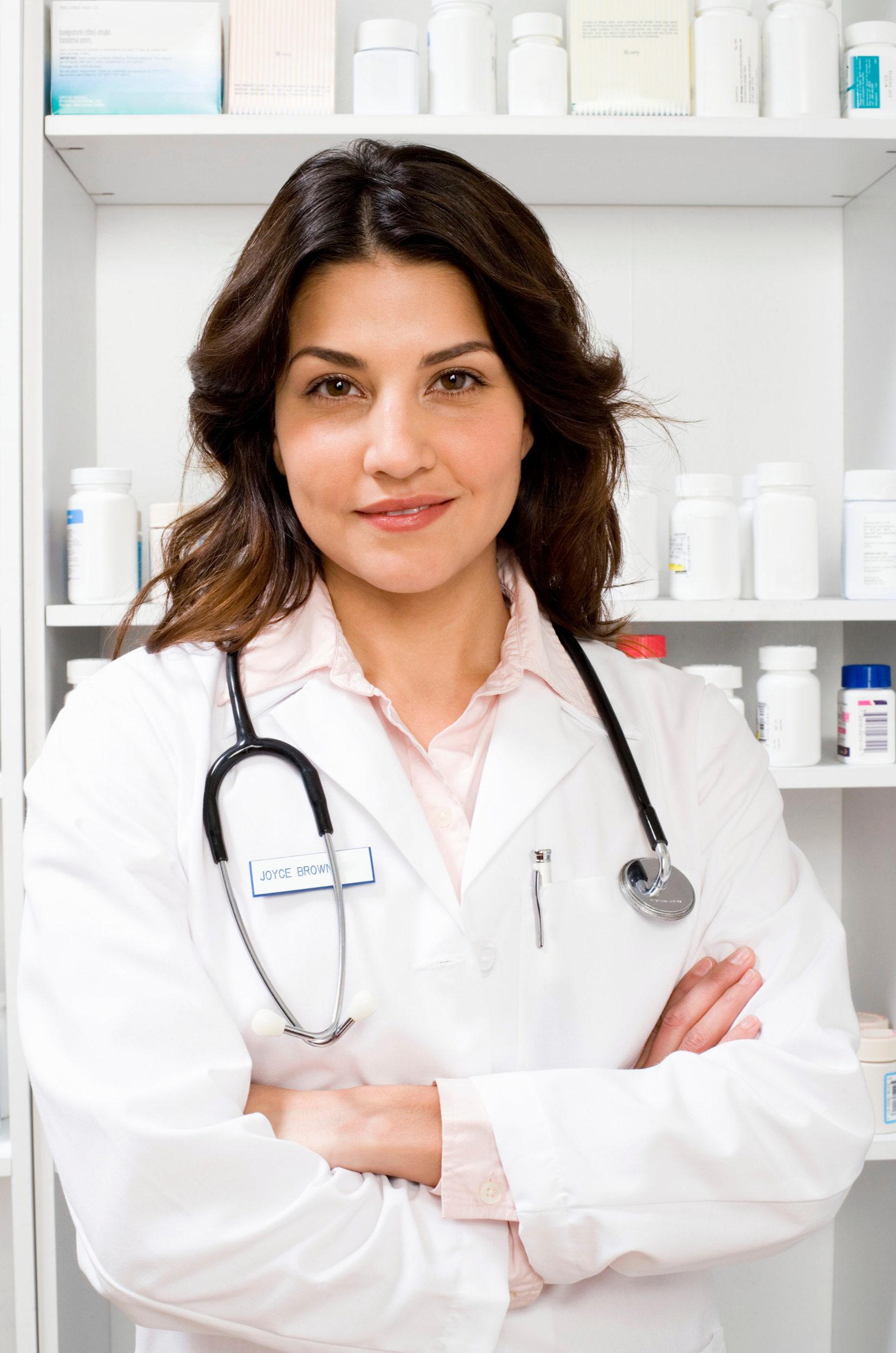 A female healthcare professional wearing a white coat with a stethoscope around her neck, standing with arms crossed in front of a medical supply cabinet.
