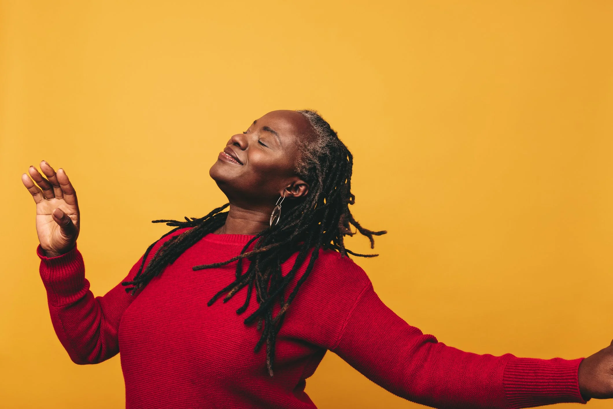 Woman with dreadlocks wearing a red sweater dancing with a joyful expression against a yellow background.