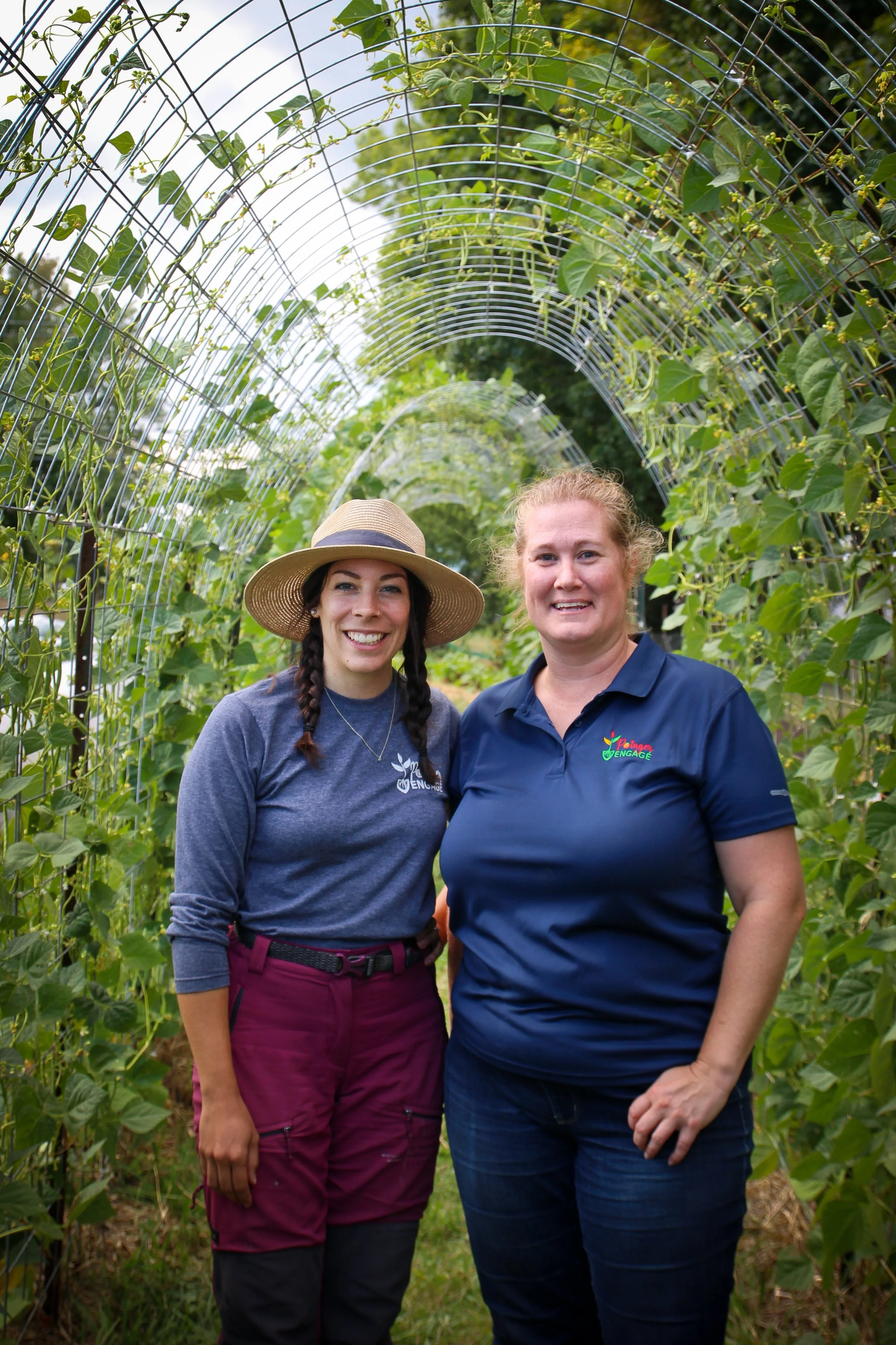 Deux femmes debout dans une serre ou un tunnel de culture, entourées de plantes vertes grimpantes, souriantes, portant des vêtements de travail et un chapeau dans l'un des cas.