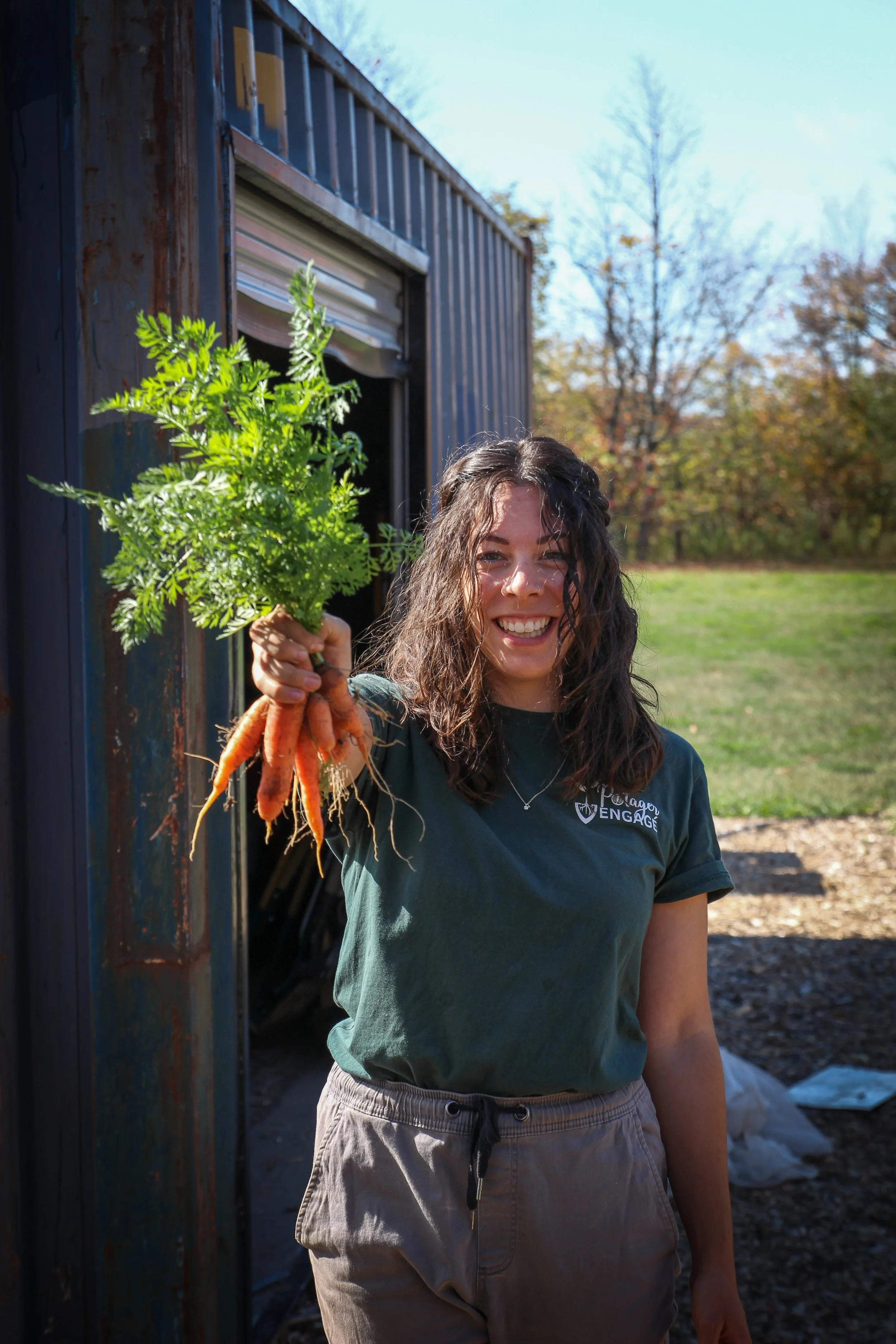 Jeune femme tenant un bouquet de carottes fraîchement récoltées devant une petite ferme ou une serre en plein air, avec un fond de ciel bleu et des arbres en automne.