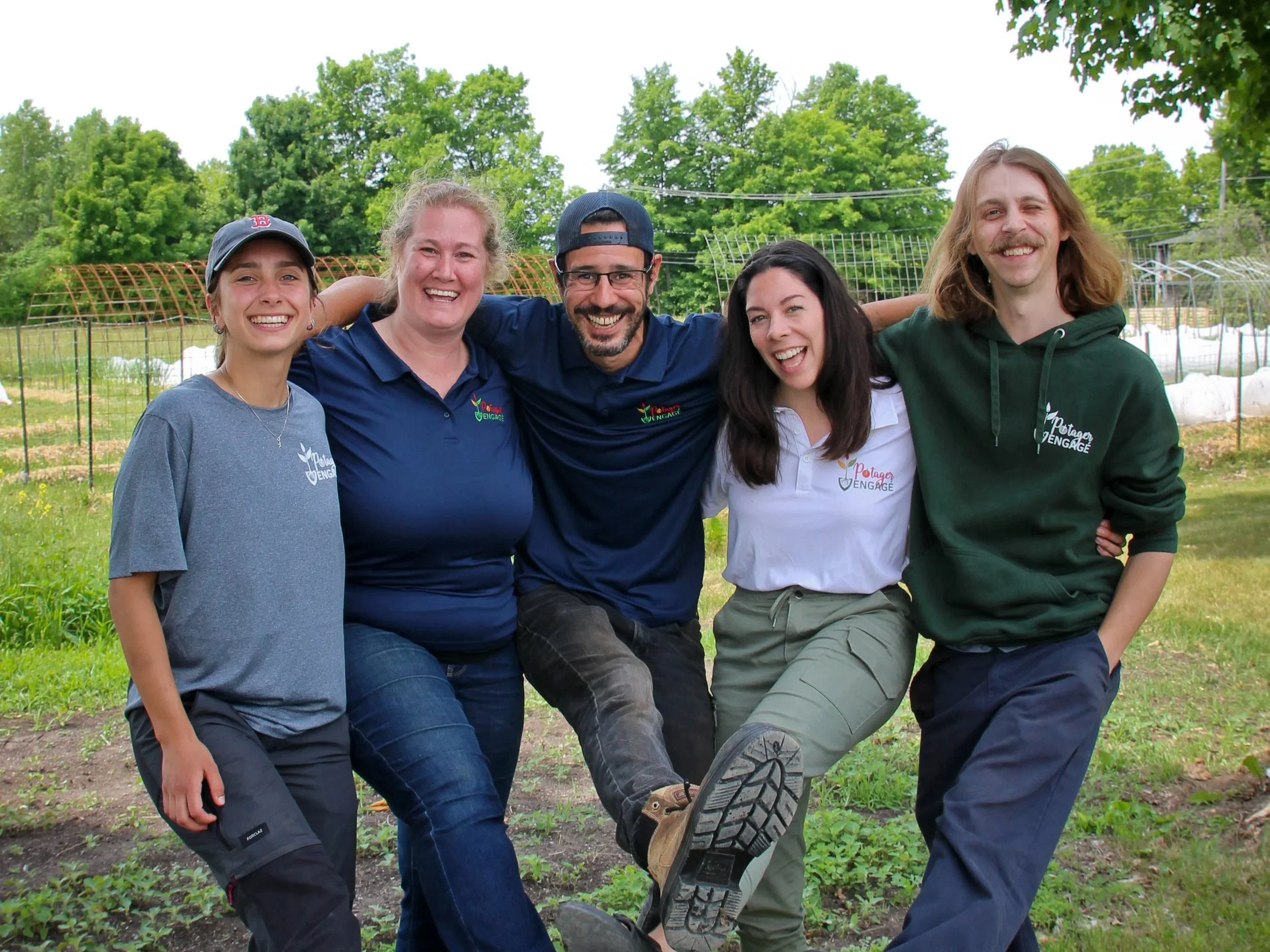 Cinq personnes souriantes posent ensemble dans un champ vert avec des arbres en arrière-plan, en lien avec un programme d'engagement communautaire.