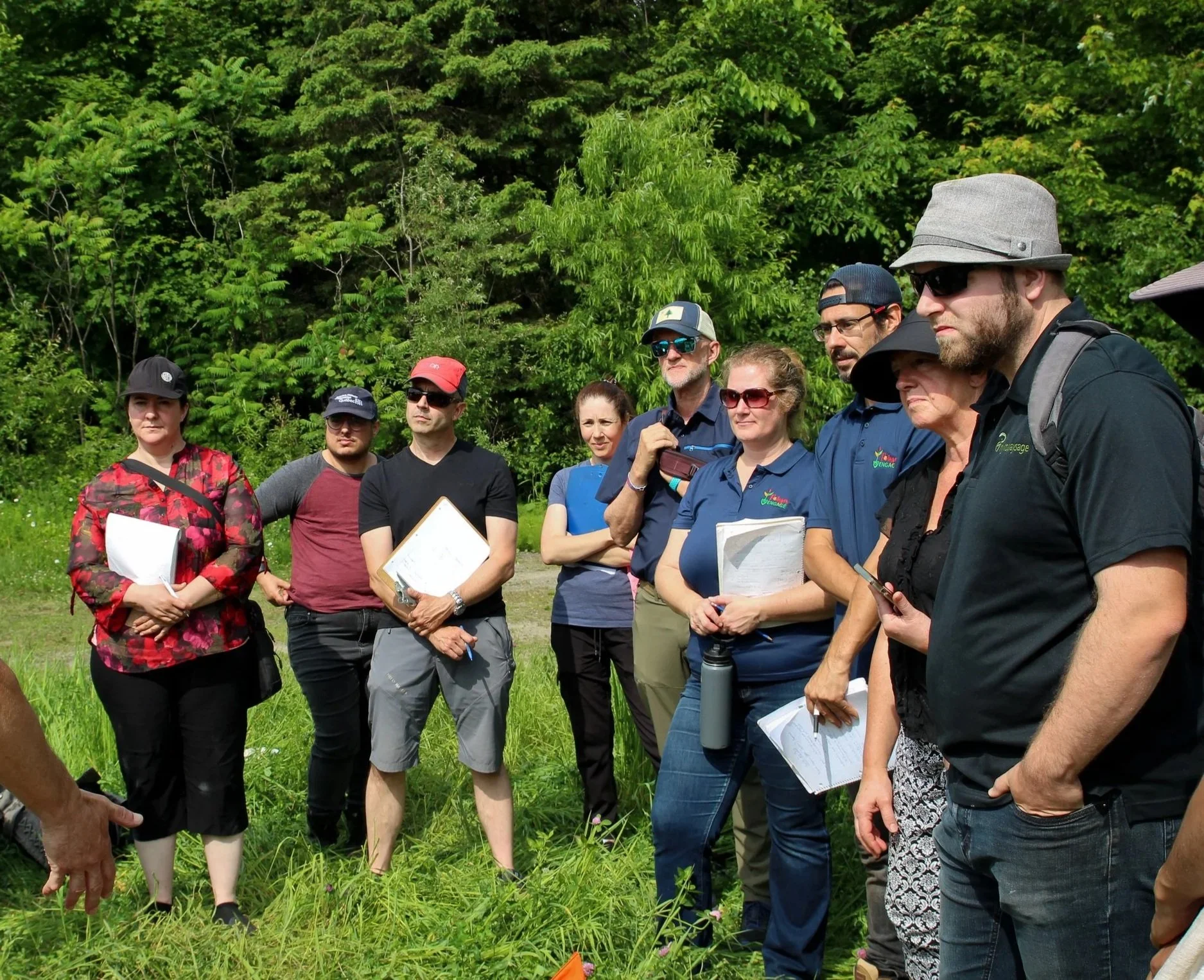Groupe de personnes debout, en demi cercle, certains tenant des carnets , des calepins ou téléphones, sous un ciel ensoleillé avec la lisière d'arbres en arrière-plan.