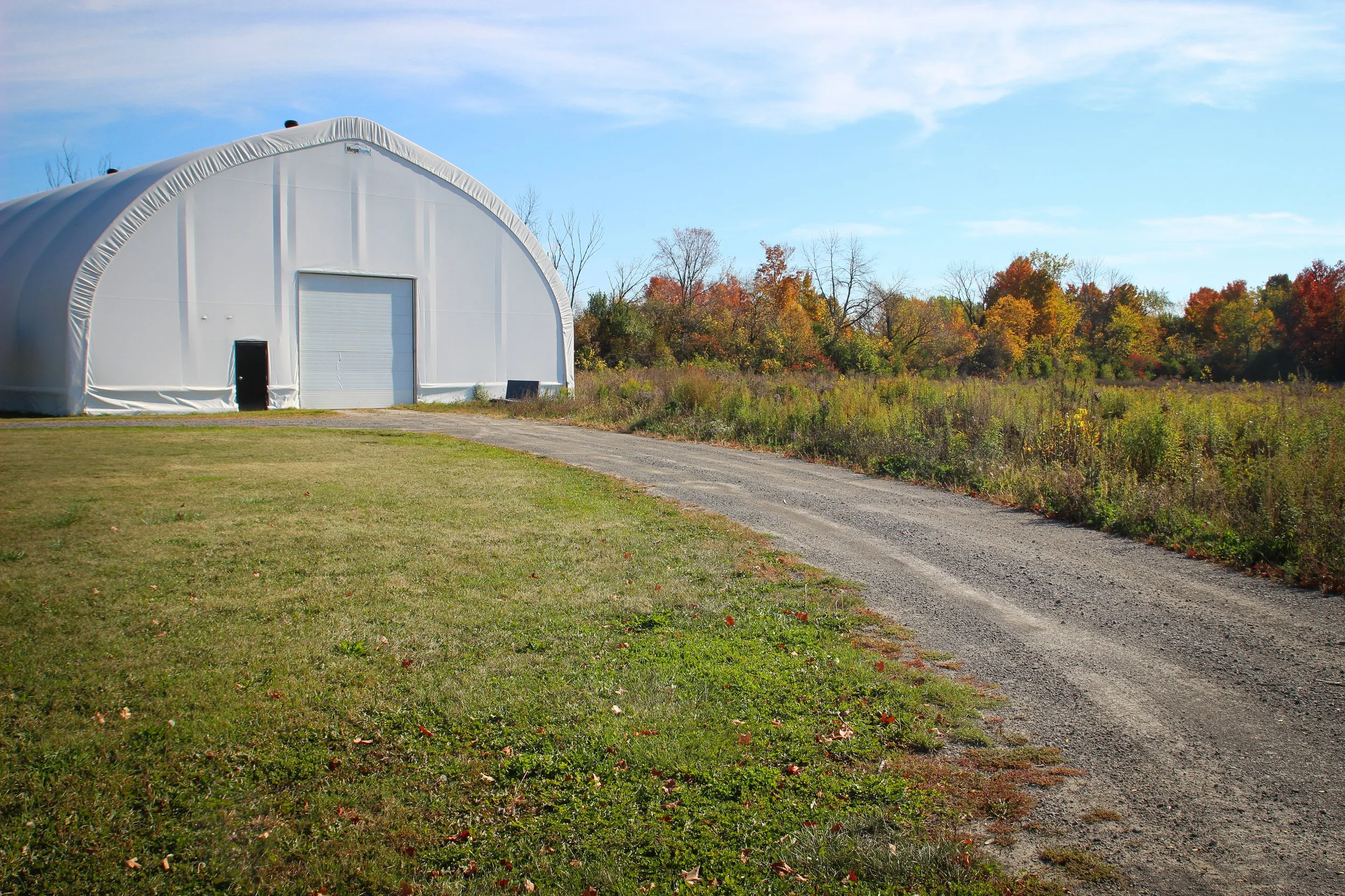 Un bâtiment en forme d'abri en toit en demi-cercle blanc, situé à côté d'une route en gravier qui serpente à travers un champ avec des arbres aux feuilles d'automne colorées en arrière-plan, sous un ciel bleu avec quelques nuages.