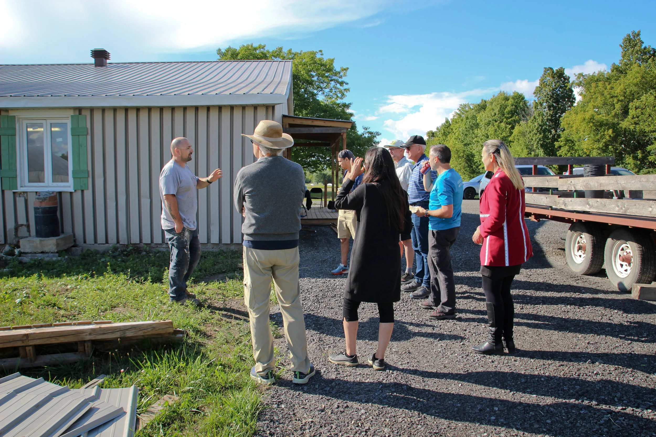Groupe de personnes en extérieur, écoutant un homme parler, devant une maison en métal, en journée ensoleillée.