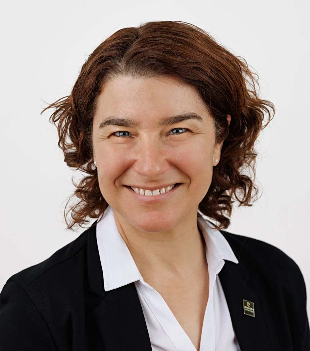 Portrait d'une femme souriante avec des cheveux courts bruns, portant un costume noir avec une chemise blanche.