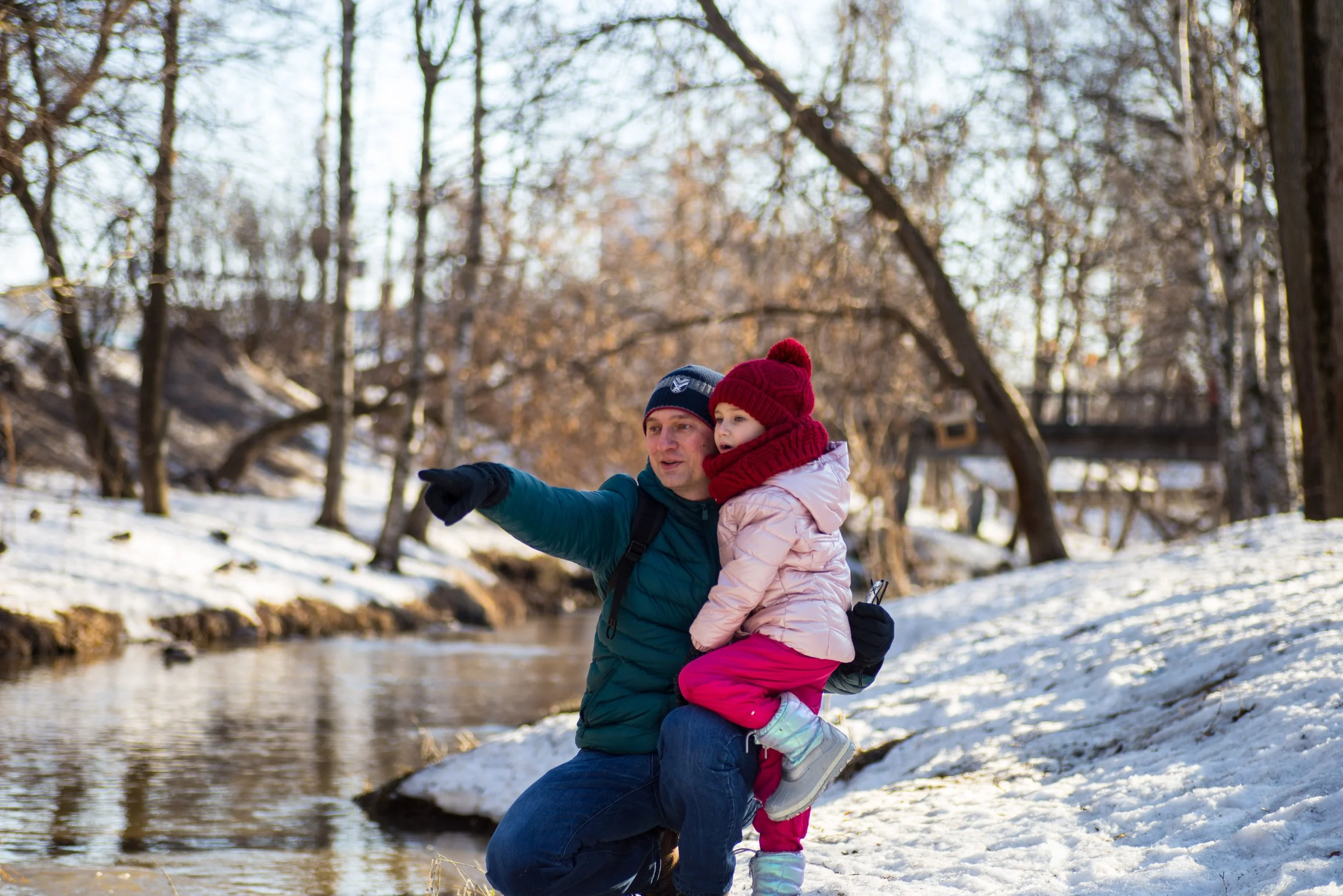 This is a photo of two family members outside. An adult is crouched down beside a river, in the snow, showing a child something in the distance.