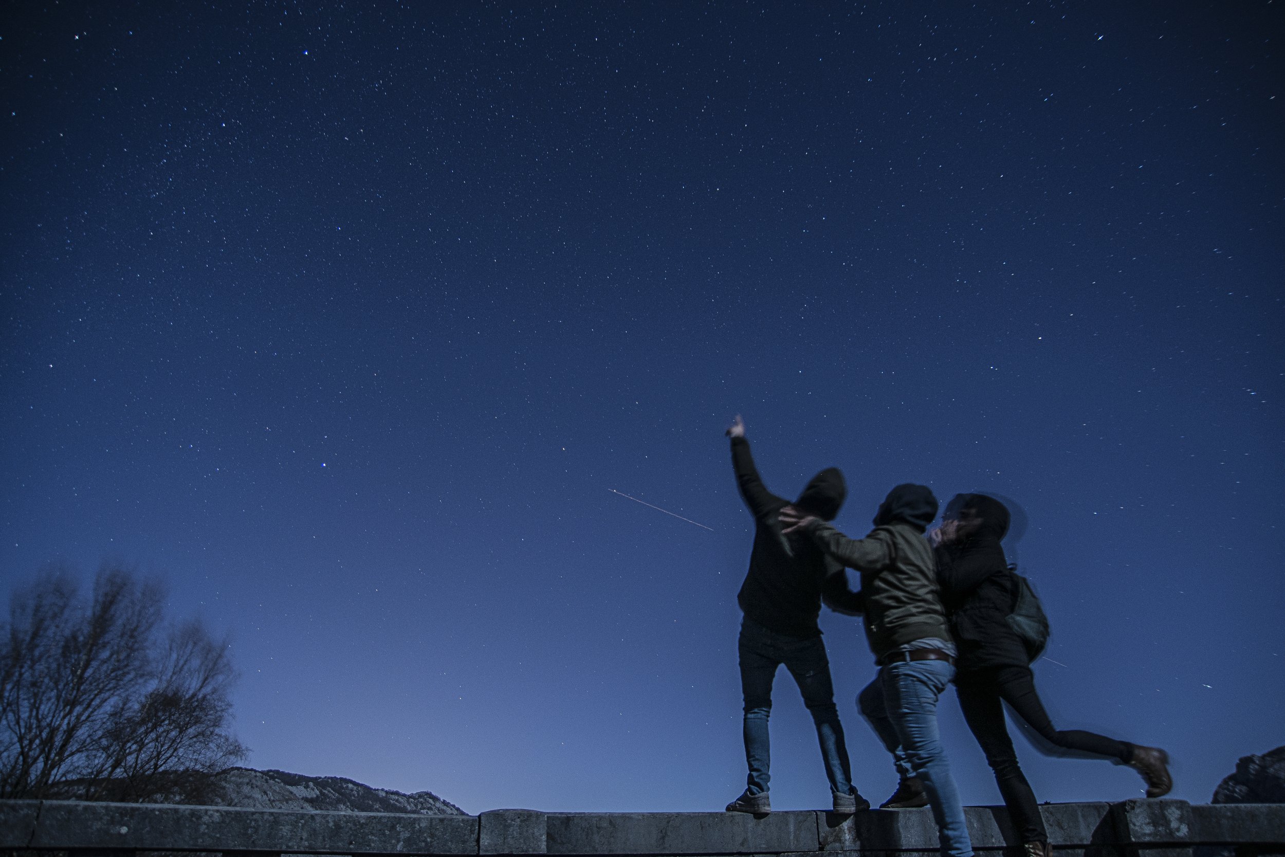 This image is of a family looking up at the stars together, under a night sky. Three members of a family are standing close to each other, with one family member pointing to the stars.