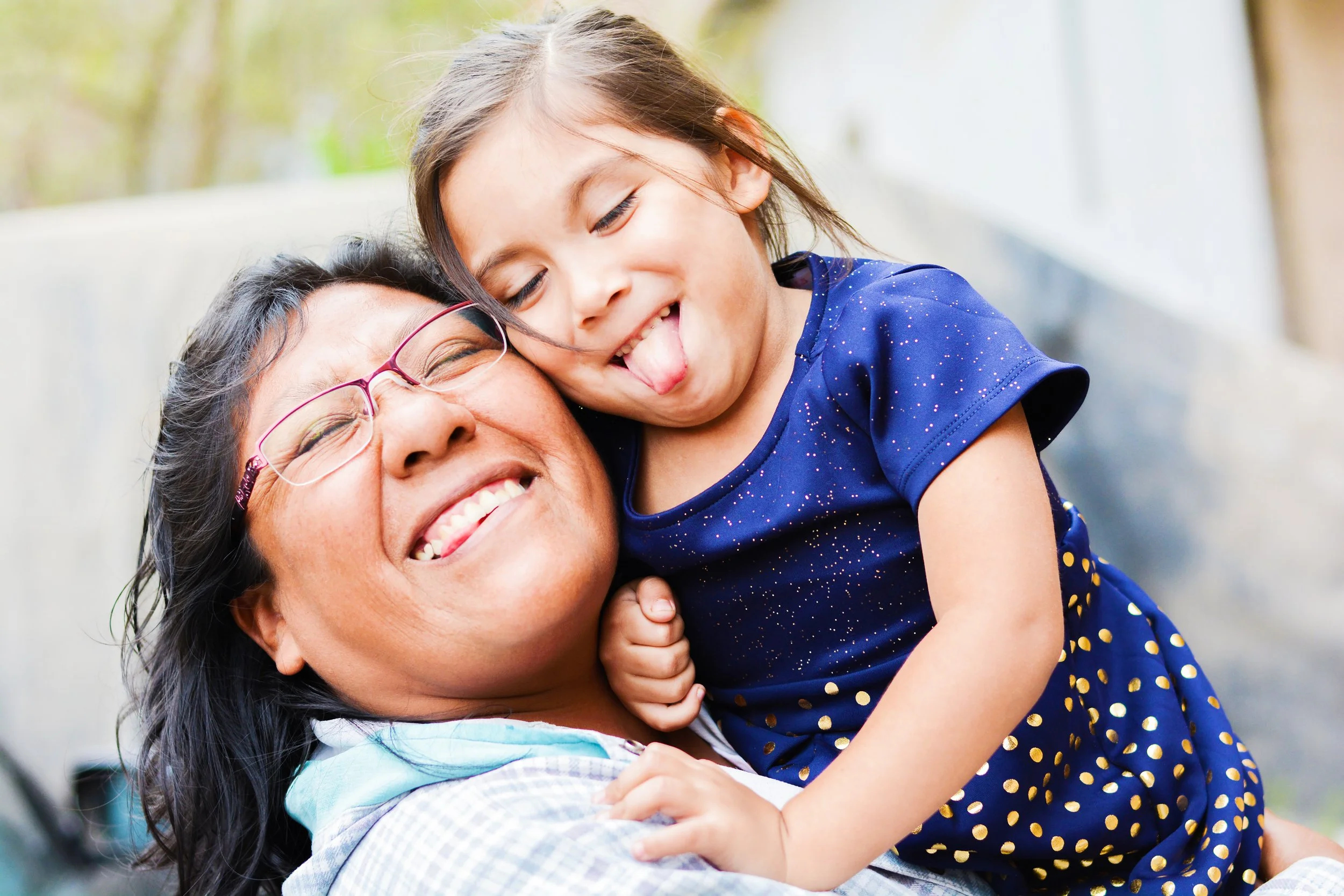 This image is of two family members being silly together. An adult is holding a child close as they smile, laugh, and stick out their tongues in fun.