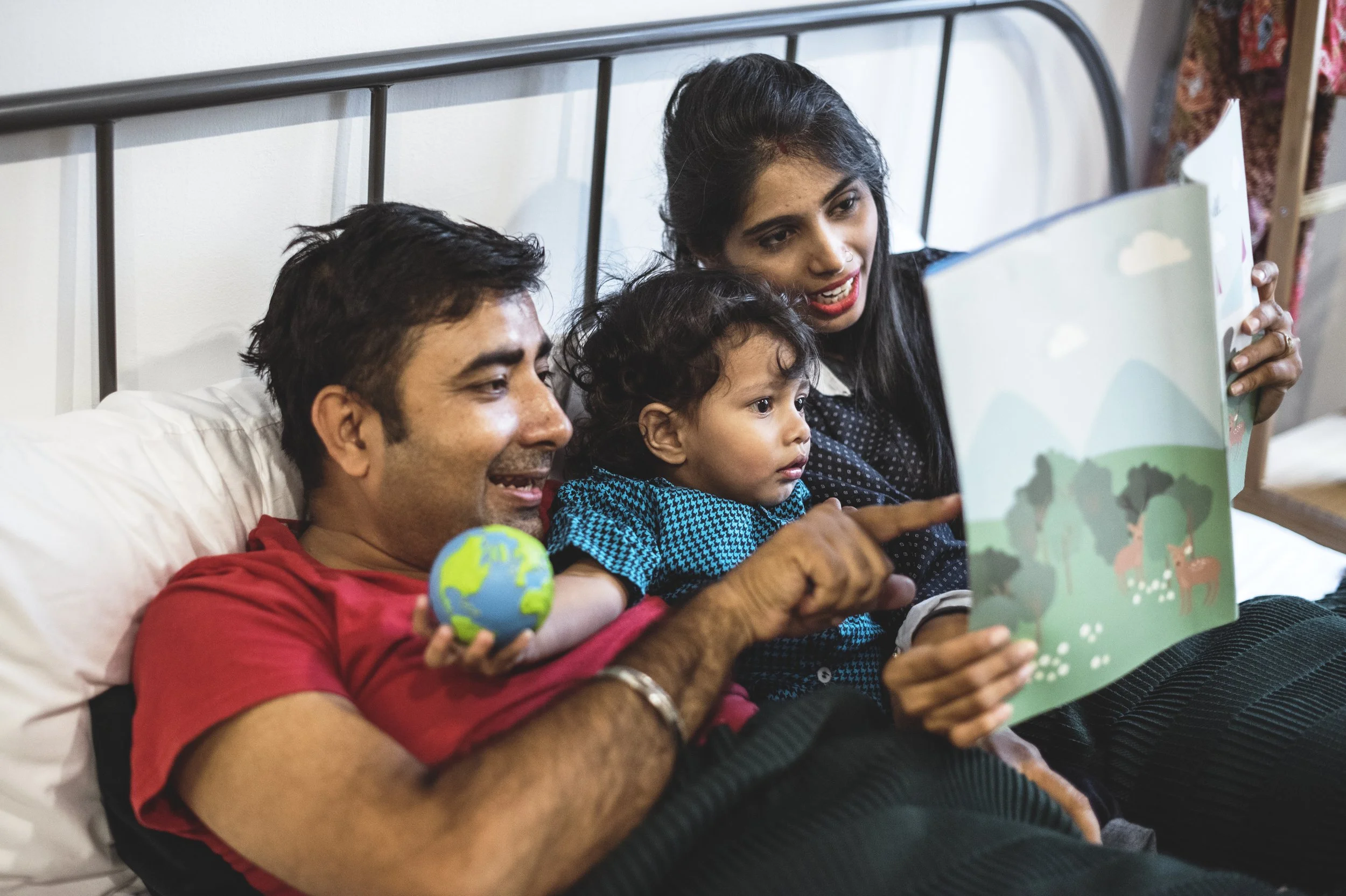 This is a photo of three family members, lying in bed and reading a book together. Two adults are reading a book to a small child, and pointing at the book to show the letters and pictures.