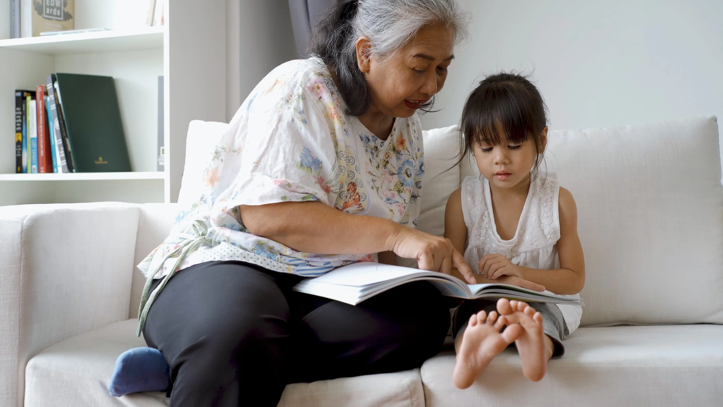 This is a photo of two family members, sitting on a sofa and reading a book together. There is an older adult pointing to a book that is on their lap and the lap of the child beside them. They are both very interested in what the book says.