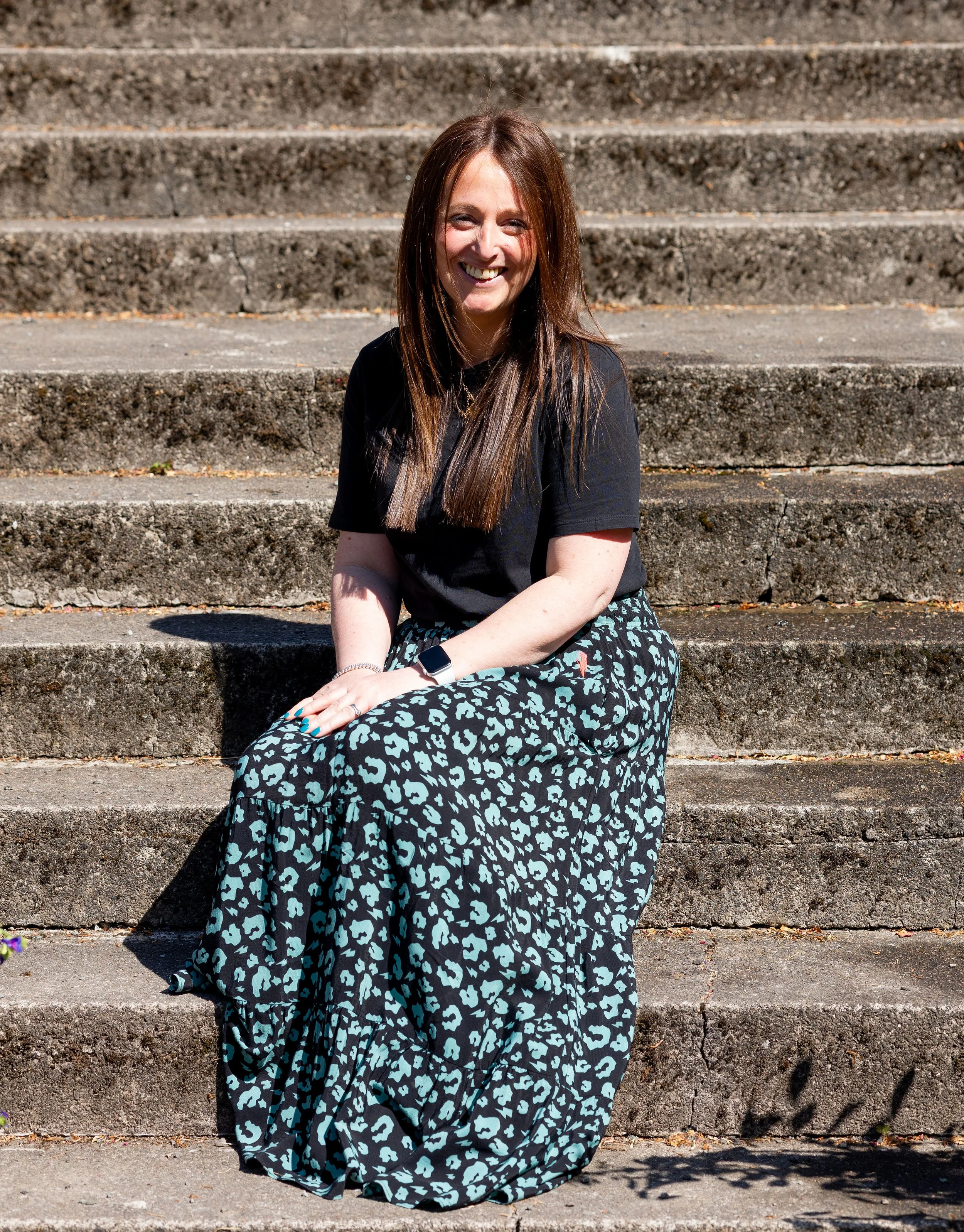Elizabeth, the calligrapher and owner seated at the bottom of some stone steps, wearing long brown hair over shoulders, a black tshirt and a green and black full length skirt