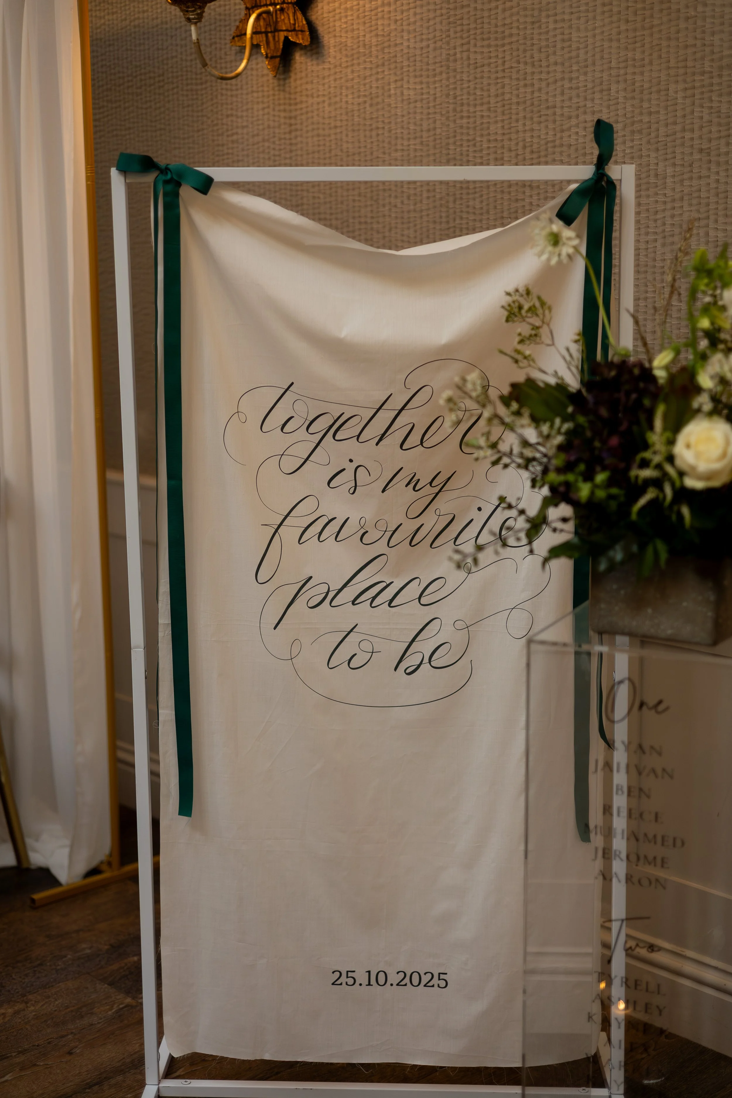 a fabric wedding banner hung inside a wedding venue with dark green ribbons and bows. The banner has calligraphy reading together is my favourite place to be. You can see a floral arrangement in the foreground