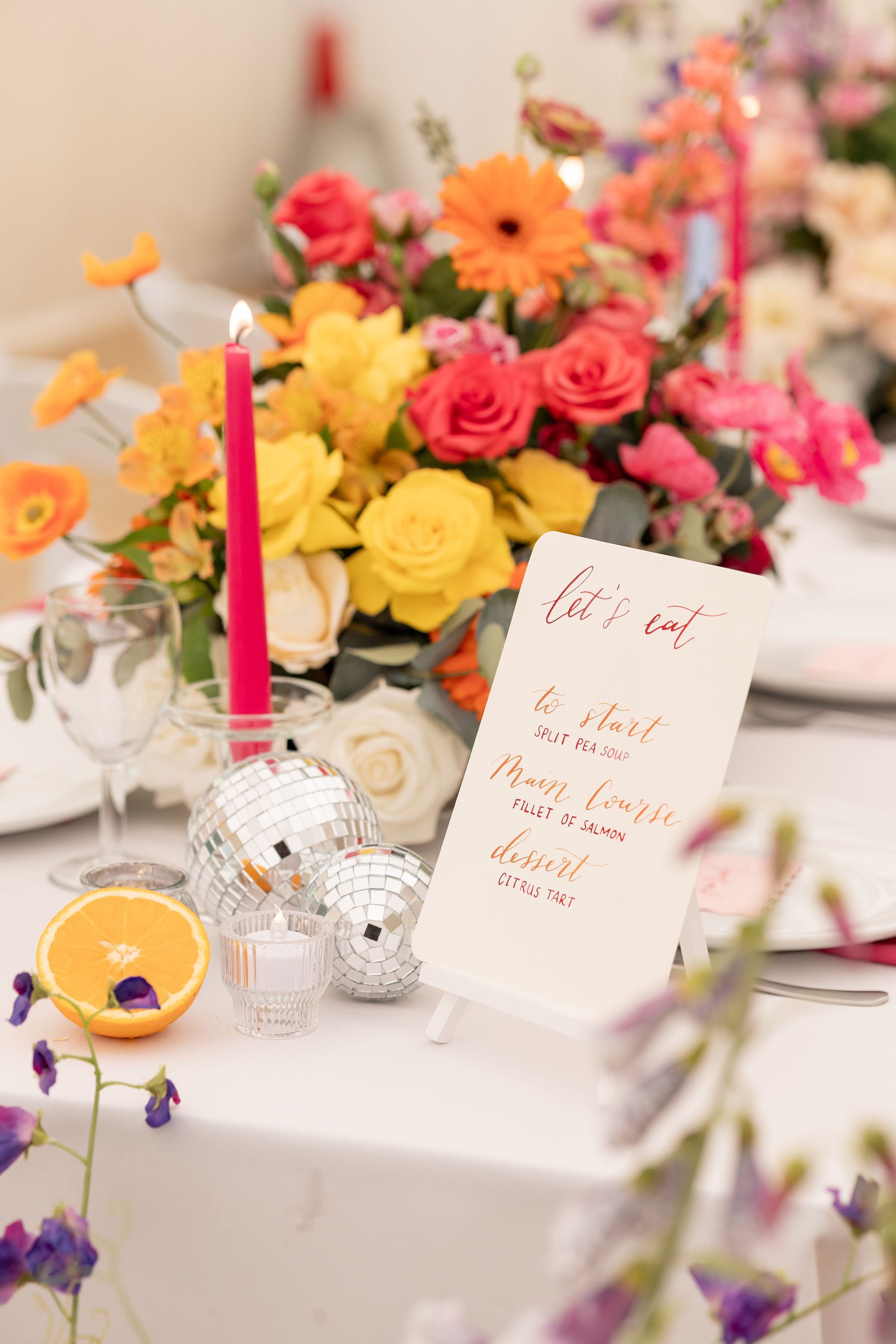 A colorful centerpieces with pink, orange, yellow, and white flowers, a pink candle, a half orange, small candles, and disco balls on a table at an event. A menu sign includes courses like split pea soup, salmon fillet, and citrus tart.