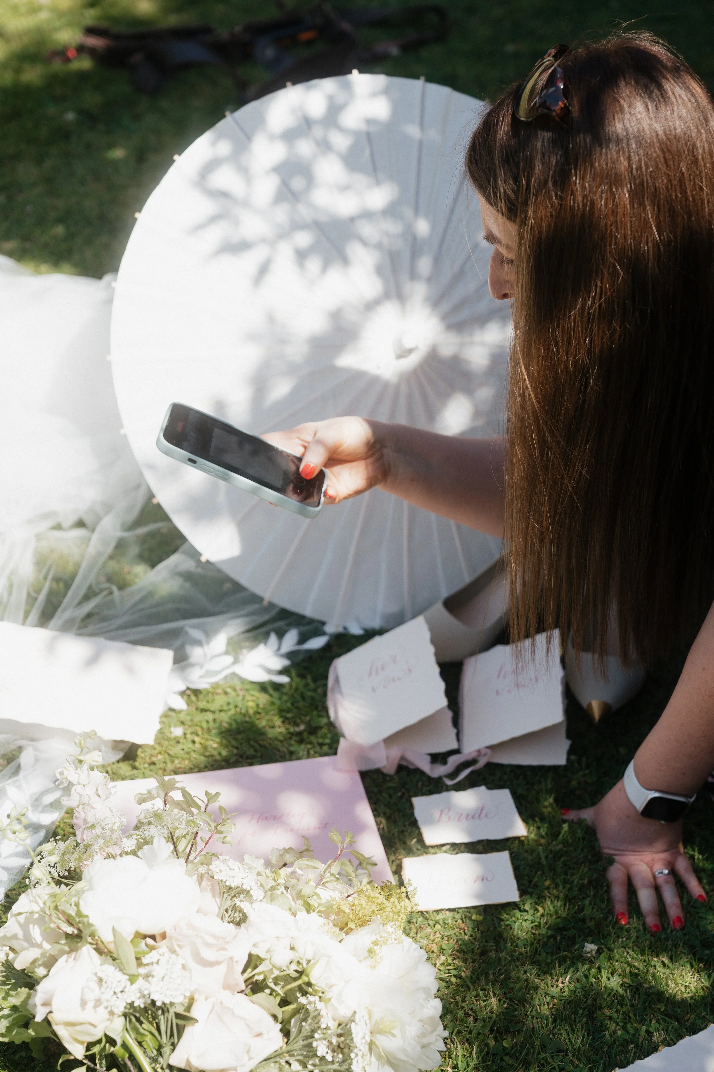 vanessa pic of elizabeth bending over flat flay on the grass with invitations, wedding flowers, parasol and veil. Elizabeth is directing her phone to the flat lay