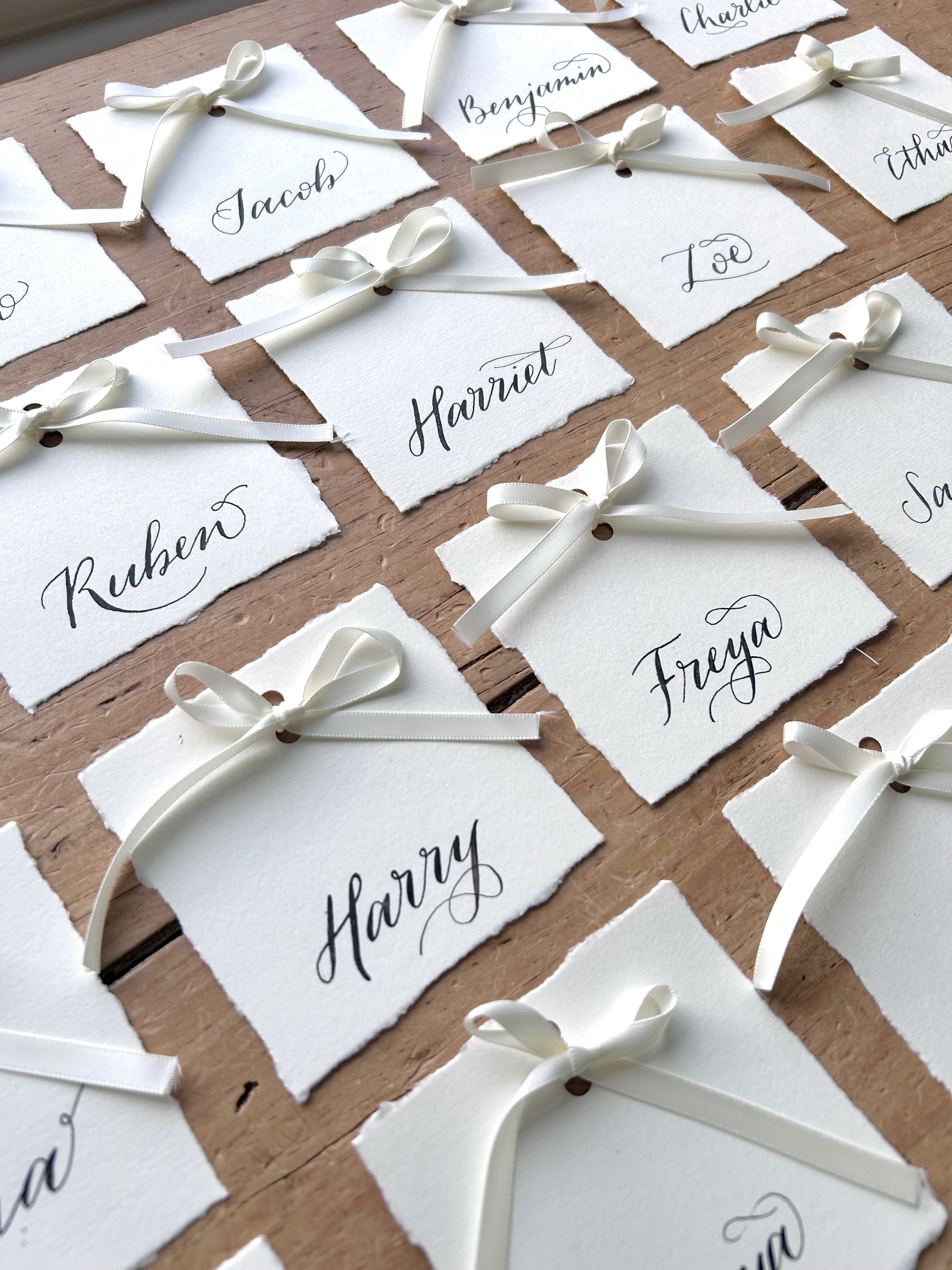 Lots of placecards lined up in rows. Cream card with hand torn edges and a delicate cream bow tied in satin ribbon. Names are hand lettered in black ink calligraphy.