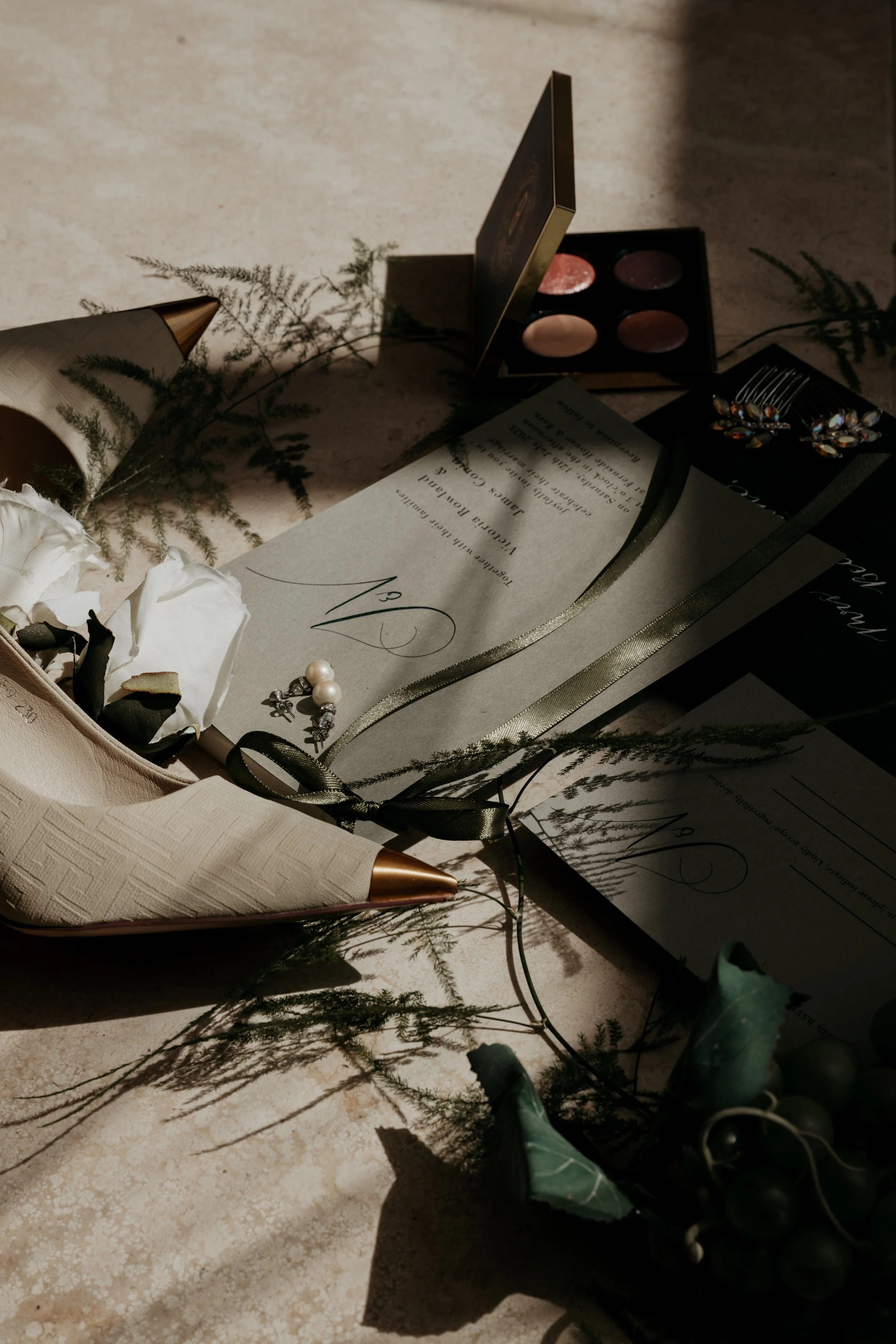 flat lay invitation in sunlight with make up pallate, pointed shoes and ferns