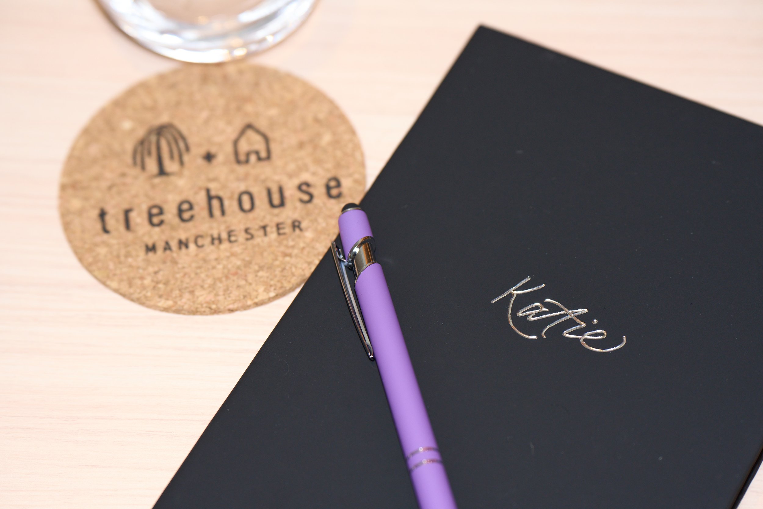 A personalised black notebook with the name Katie foiled in calligraphy. A lilac pen is resting on the notebook and a coaster for Treehouse Manchester Hotel can be seen next to it