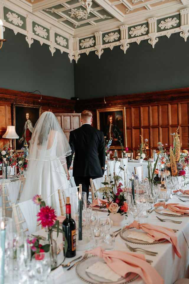 Couple in wedding attire standing in an elegant decorated dining room with a long table set for a formal event, adorned with floral arrangements, candles, and wine bottles, with dark green walls and framed portraits on the wooden-paneled walls.