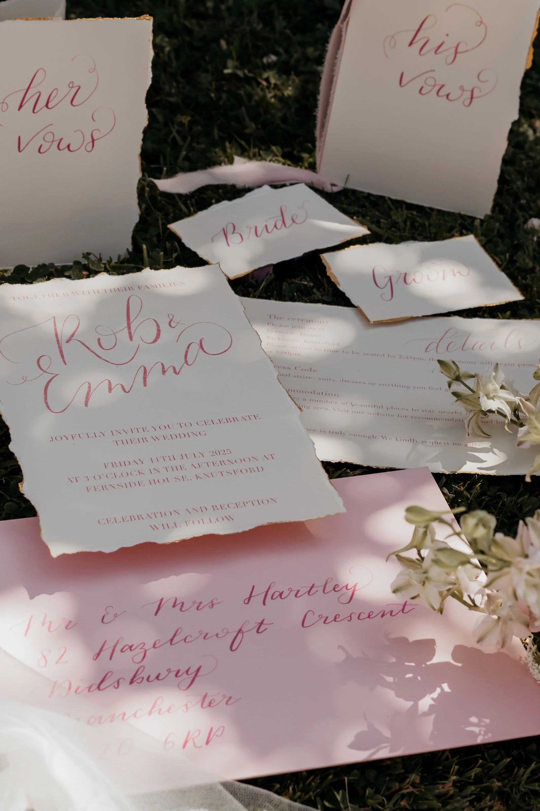 Wedding invitation and stationery items scattered on grass, including an invitation for Rob and Emma's wedding, address card, and wedding roles like bride, groom, her vows, his vows, and dearest. There are also floral decorations.