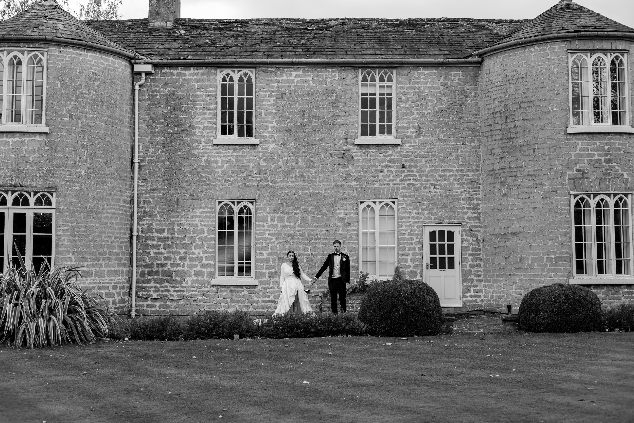 a black and white image with a country house wedding venue and a bride and groom looking at the camera. the bride and groom are holding hands
