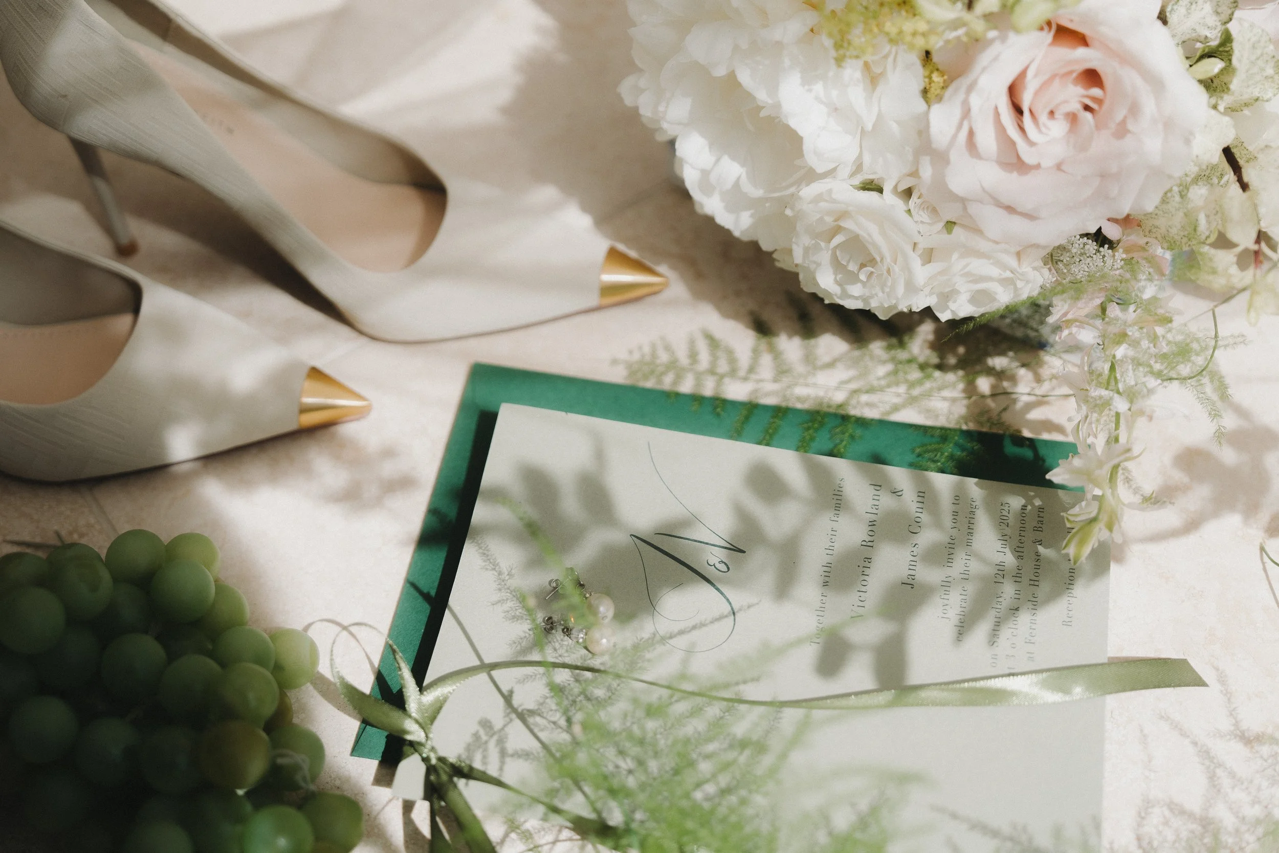 Wedding invitation surrounded by white and pale pink flowers, green grapes, and a pair of white high-heeled shoes with gold tips on a light-colored table.