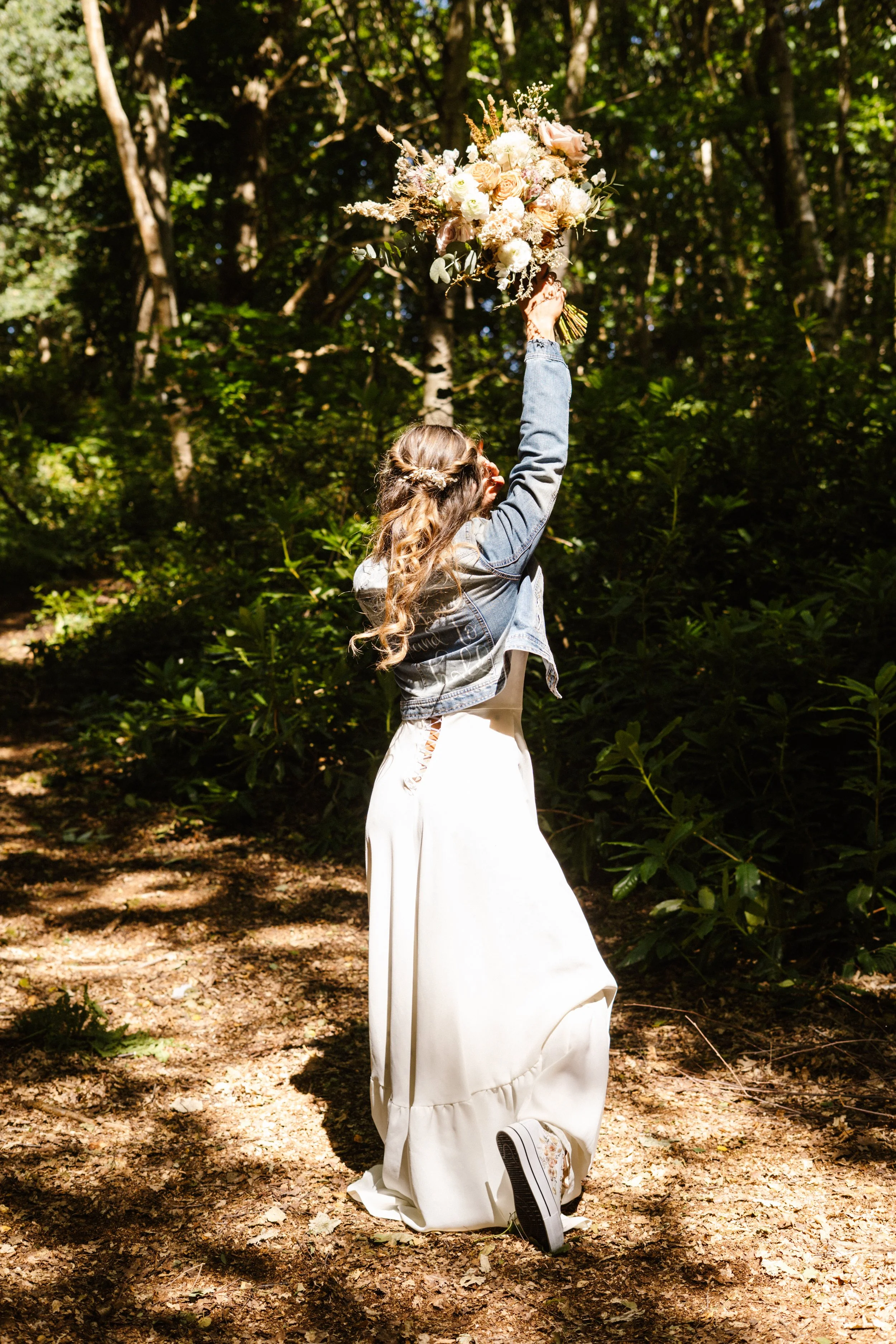 A woman in a white long skirt and denim jacket holding a large floral bouquet raised above her head in a wooded area.