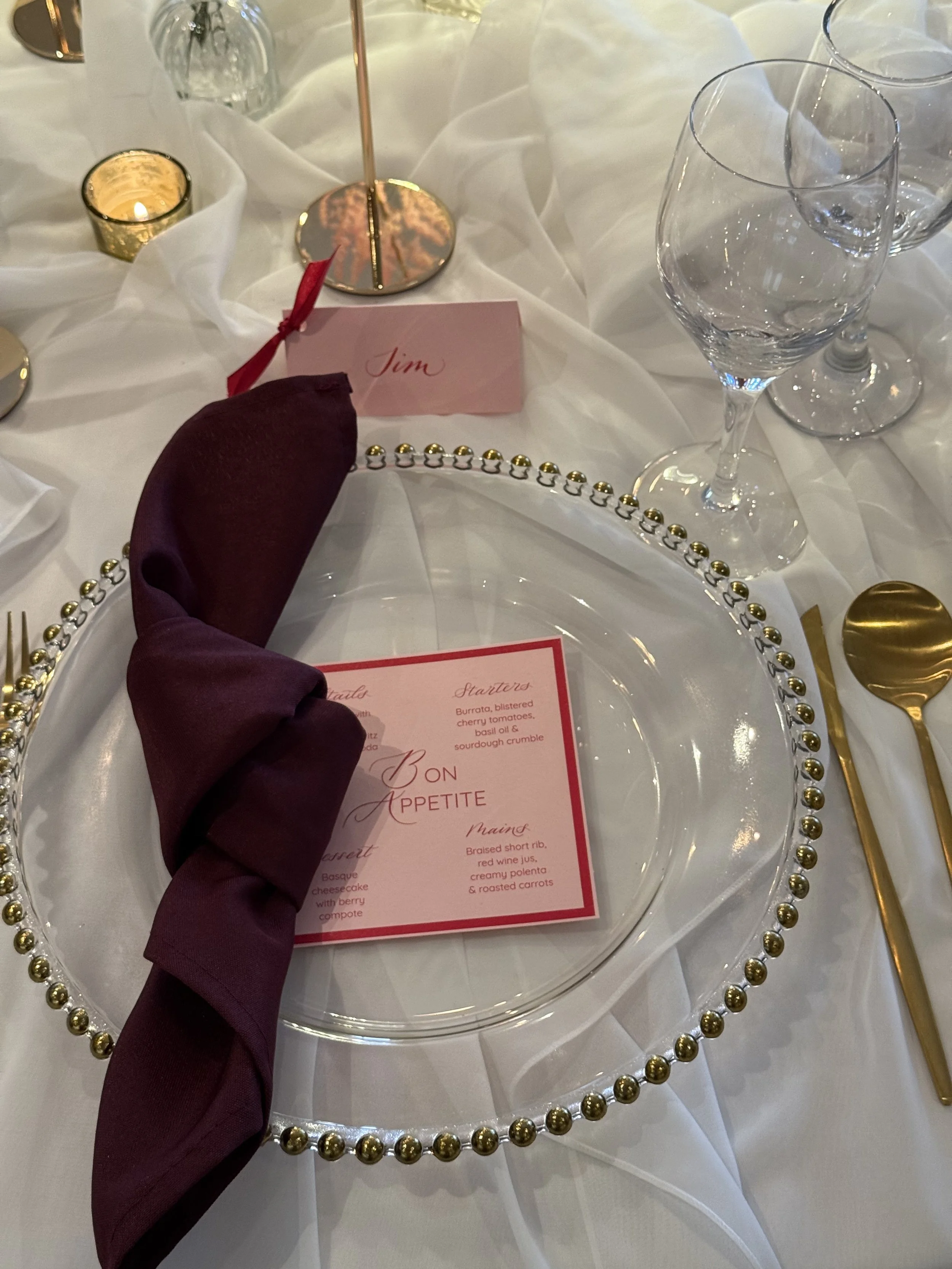 a wedding table showing candles and a white cloth and a glass charger plate. There is a pink place card and pink menu with red calligraphy ink details