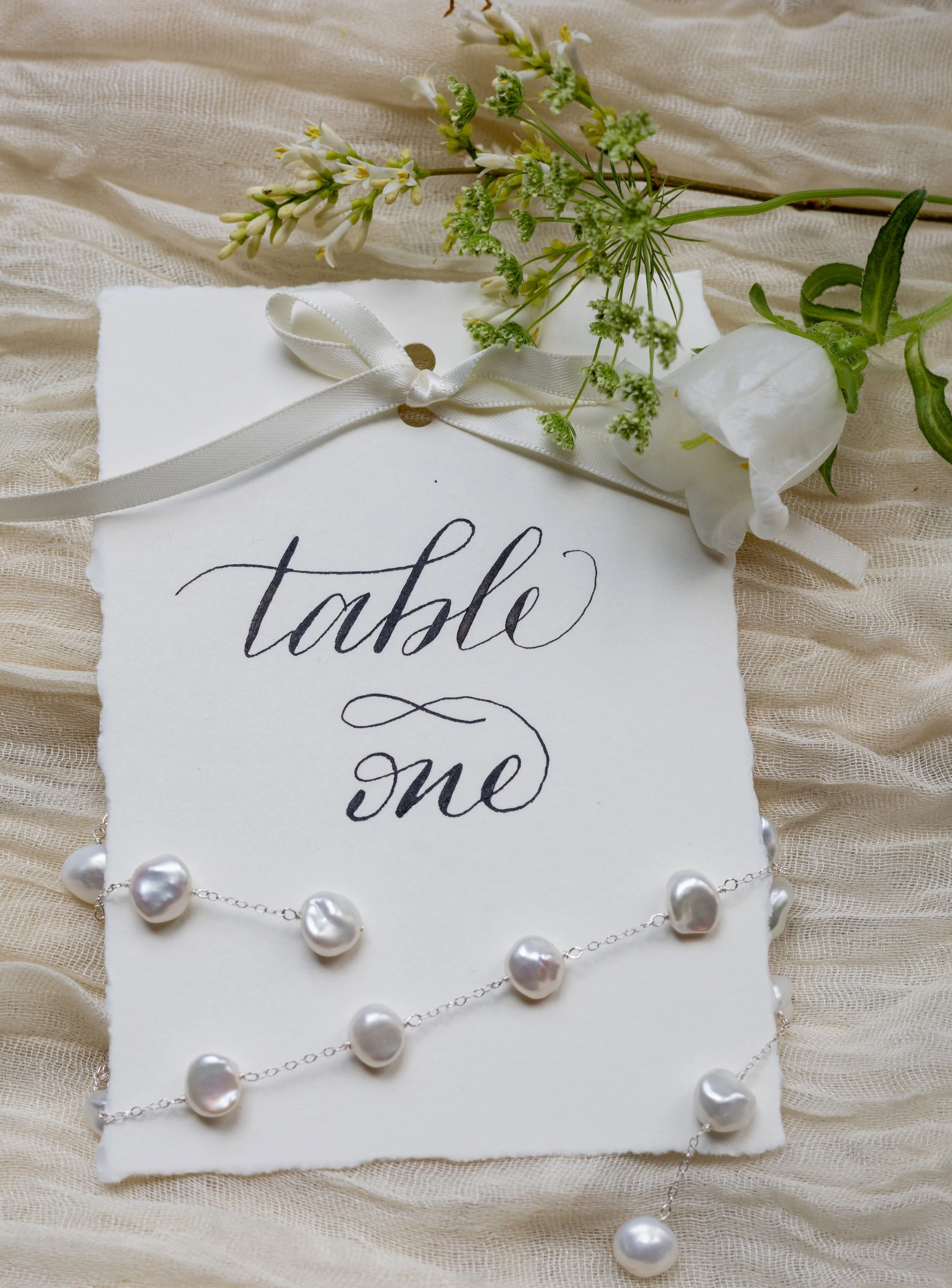a flat lay with soft chiffon featuring a calligraphy table number with hand torn edges and an elegant neatly tied bow at the top. It is draped with pearl jewwellery