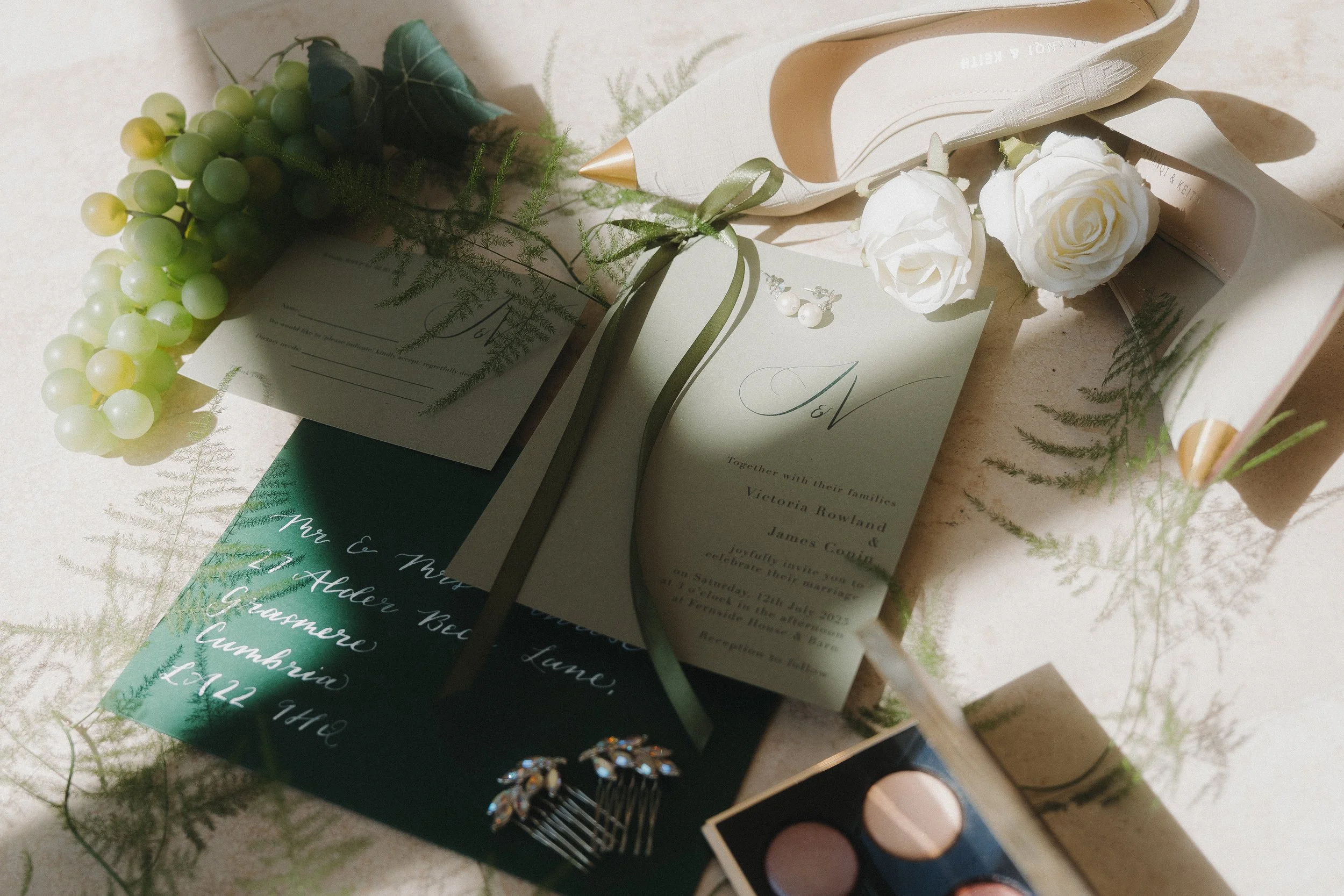 Wedding invitation suite with greenery accents, pearl earrings, a pair of white roses, a ring box with two rings, a watercolor palette, and greeting cards on a light surface.