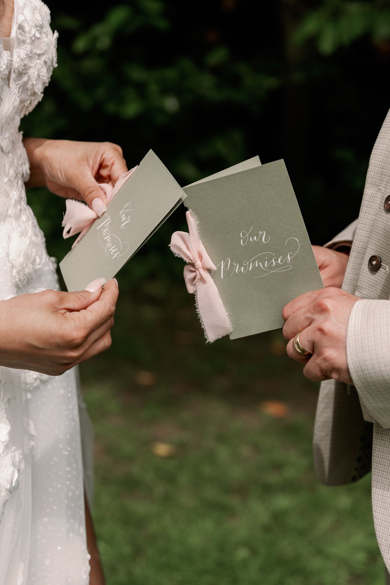 Close-up of a bride and groom exchanging vows, holding cards that say 'Our Promises' with pink ribbon accents, during a wedding ceremony outdoors.