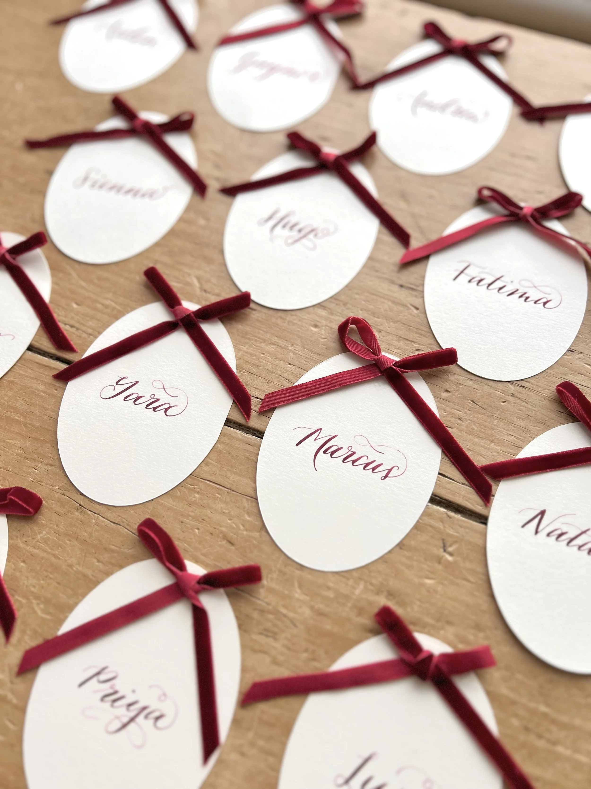 a selection of cream, ivory place cards laid on a wooden table. each place card has a name hand lettered in burgundy ink in calligraphy. each place card is finished with a burgundy narrow velvet bow