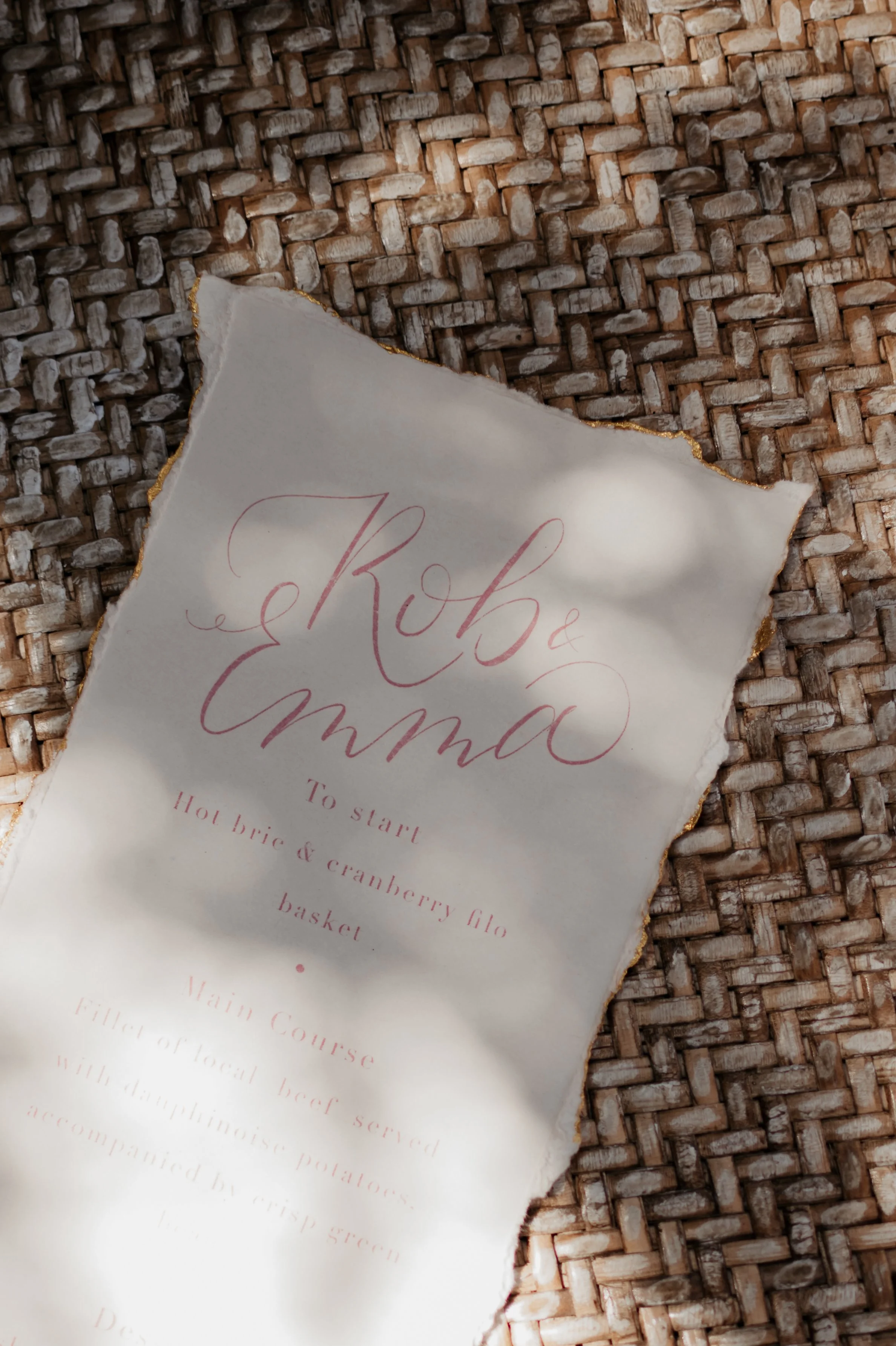 A menu with torn edges on a woven surface, featuring elegant pink cursive text listing a brunch dish and main course.