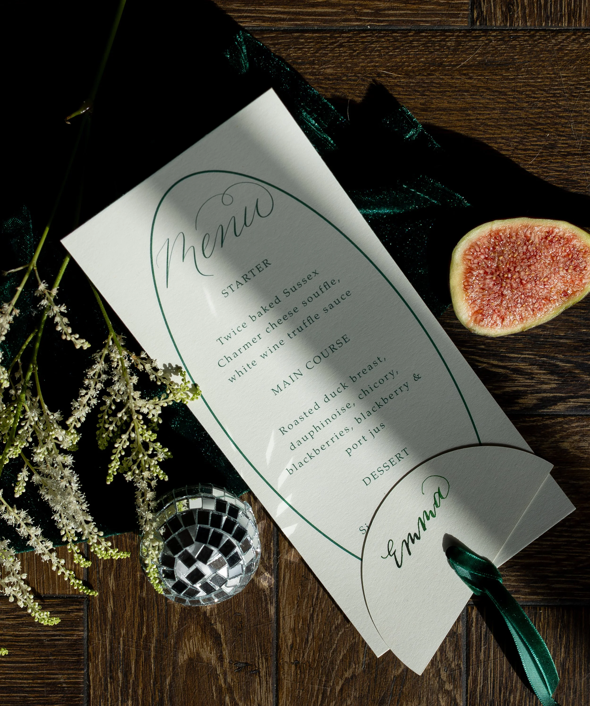 a flat lay image showing a wedding menu and attached place card (written in calligraphy with green ink). Both are attached with green velvet ribbon. The flatlay is styled with foliage, delicate ivory flowers and a mini disco ball