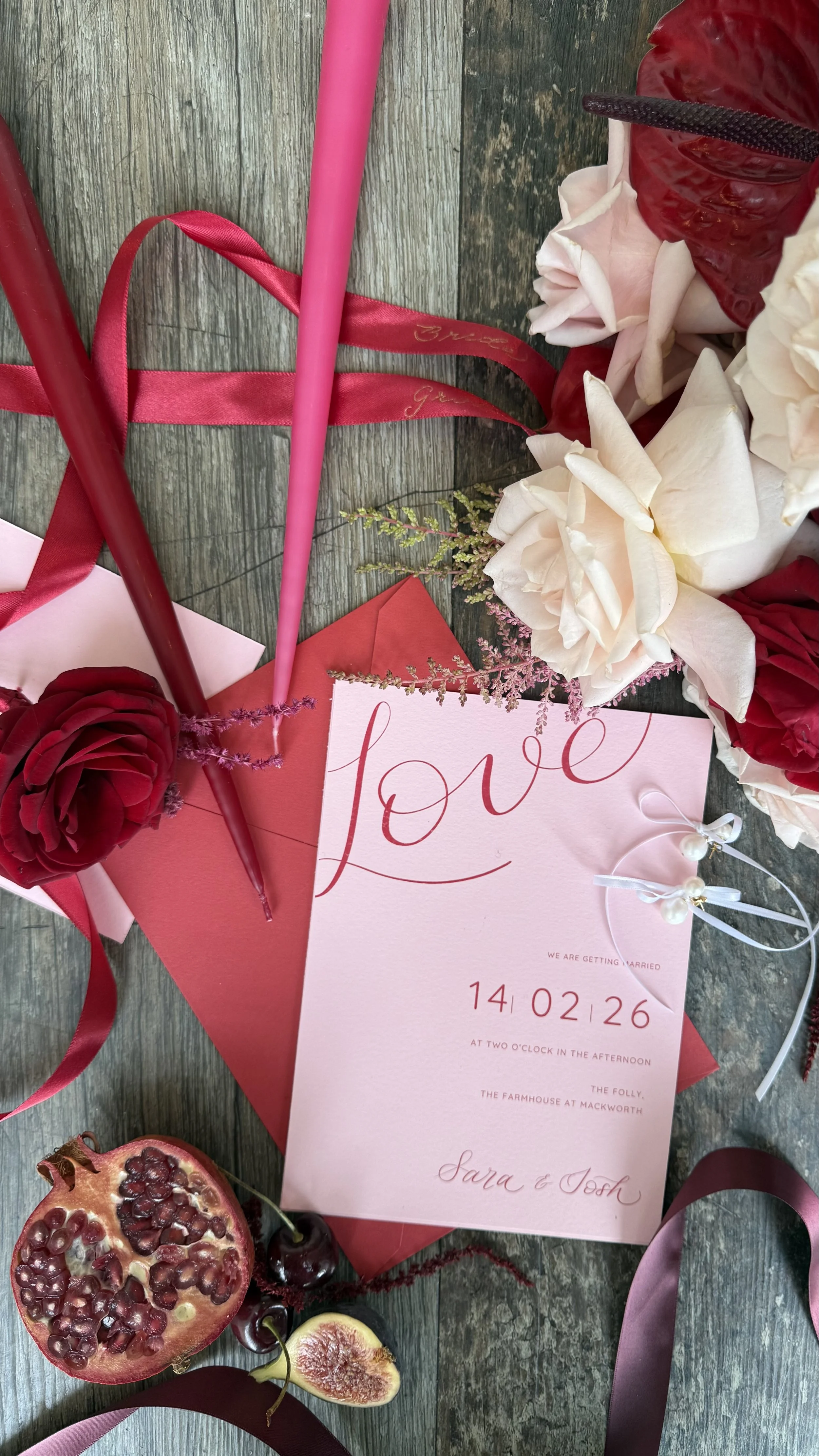 a flat lay image showing a bold pink inviatation with the word 'love' written in calligraphy. The invitation is complemented with a red envelope and pink and red florals, candles and ribbons