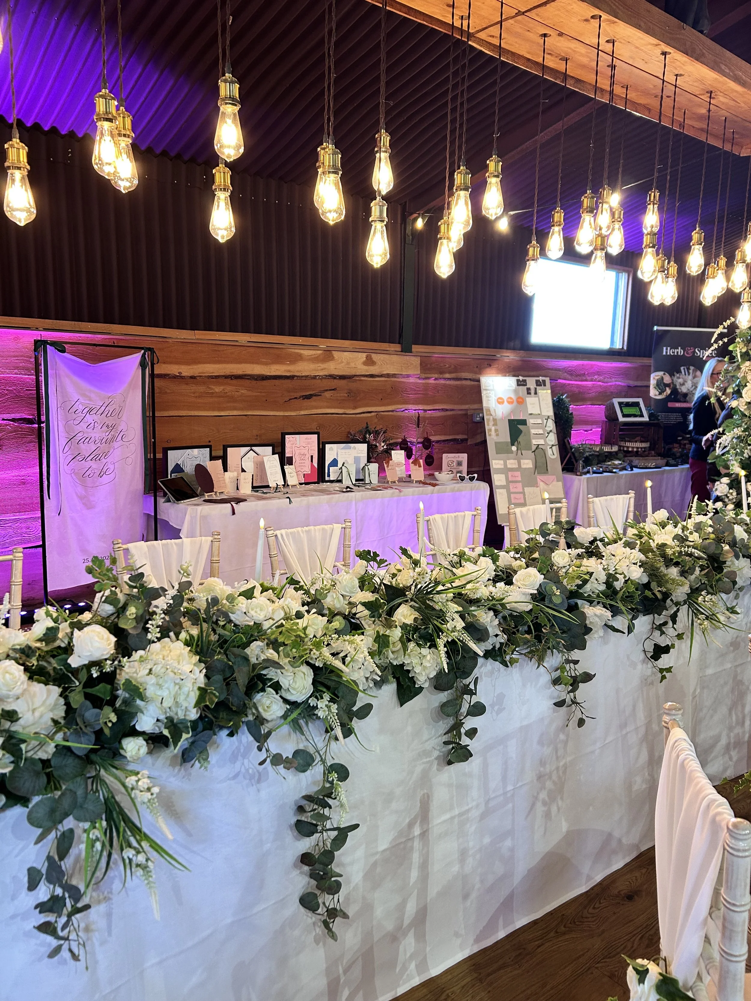 inside stock farm barn dressed with a top table and beautiful abundant floral arrangements. in the background a table can be seen showcasing stationery, invitations and signage