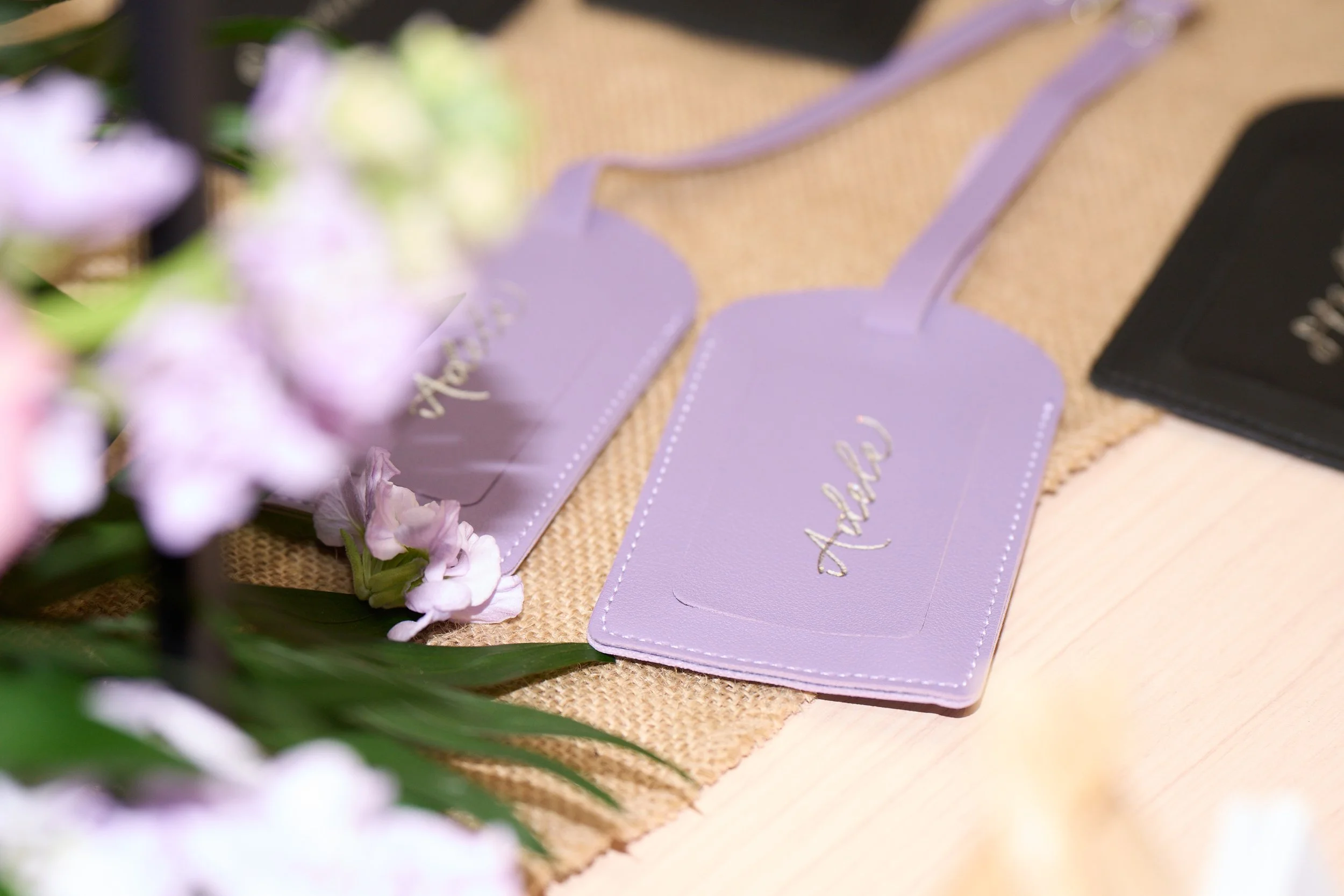 Two lilac leather luggage tags surrounded by flowers on a table. Each tag is personalised using  silver foil with the names Adele