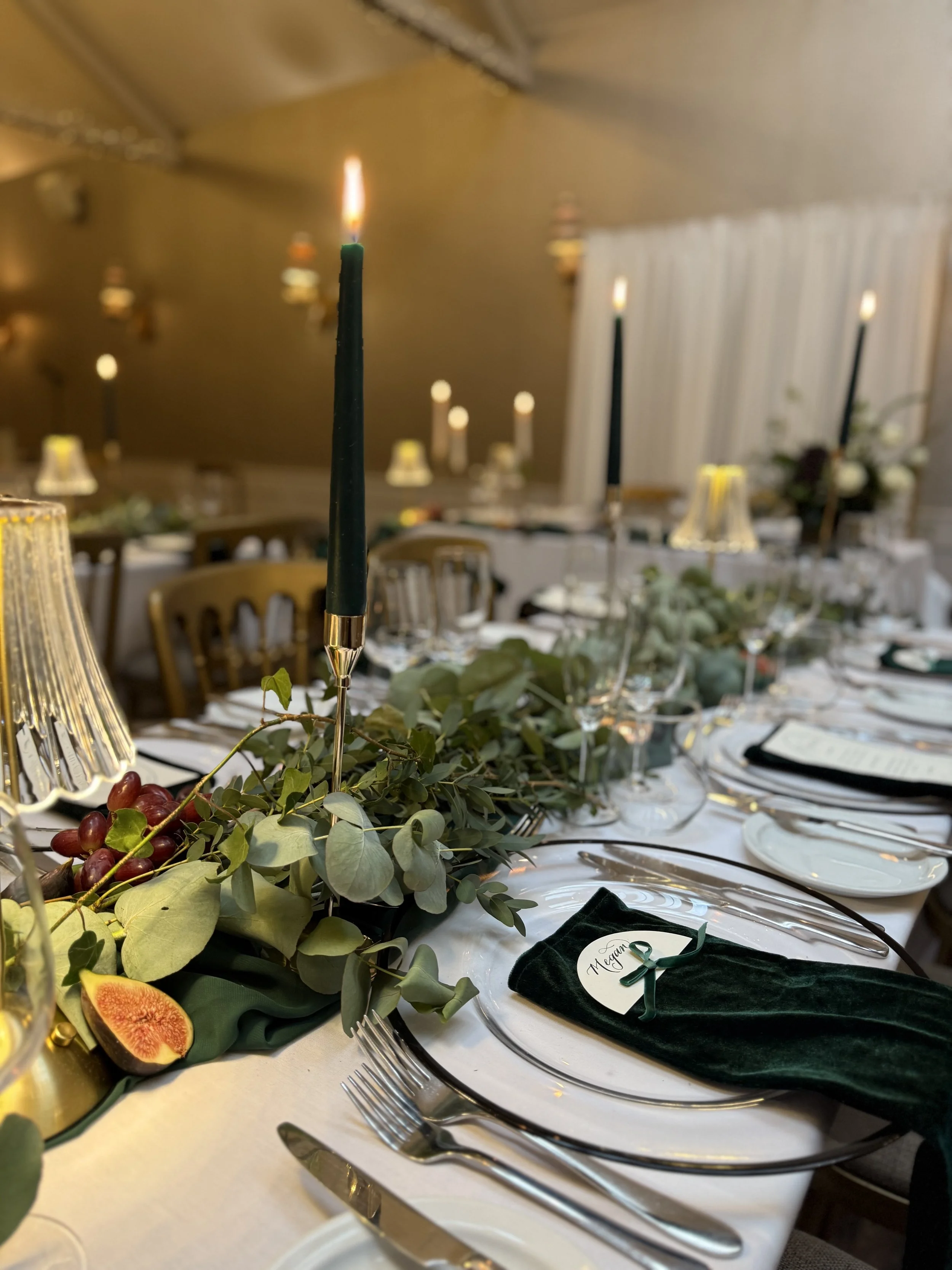 an image of a wedding breakfast table. white table cloths, dressed with green foliage down the centre, dark green tapered candles and gold, crystal lamps. you can see the gold chairs and a draped fabric backdrop behind the table