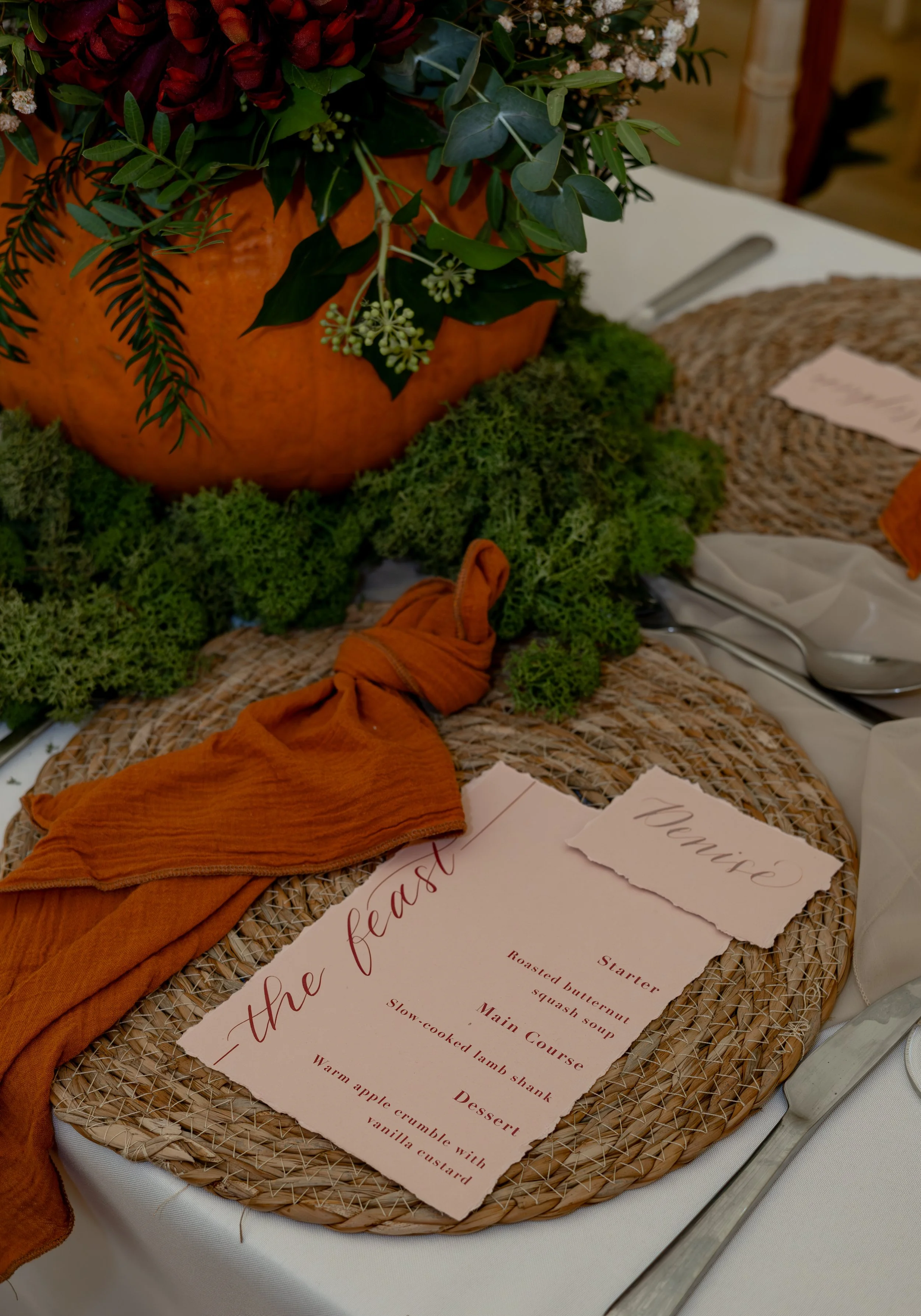 A fall-themed table setting with a decorative pumpkin centerpiece filled with greenery, surrounded by moss. There is a woven placemat, a burnt orange cloth napkin, and a menu card with a torn paper look that lists a feast menu. A handwritten name card reading 'Premiere' is placed on the placemat, with a silver spoon and fork beside a white napkin.