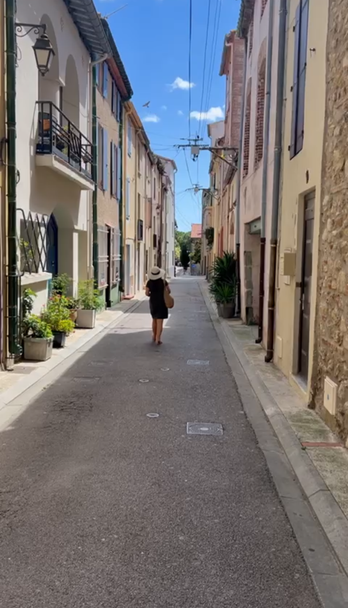A French street in Coullier with tall buildings, balconies and flowers either side. Calligrapher Elizabeth walking away from the camera showing her back and wearing a large straw hat.