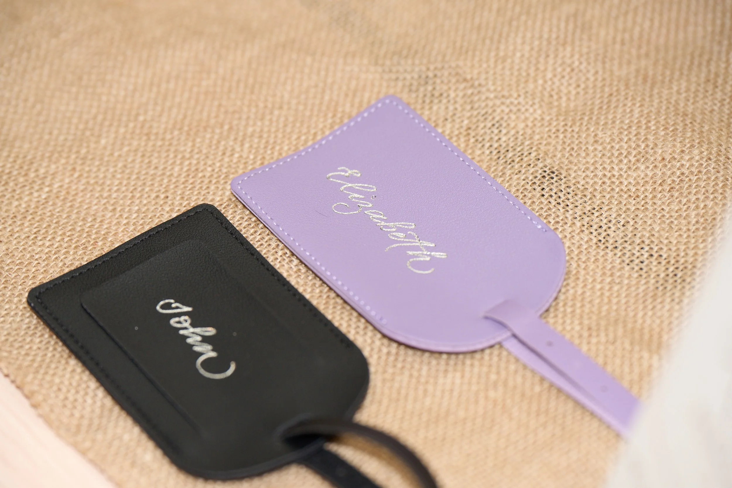 Two leather luggage tags on a hessian table cloth. Each is foiled in calligraphy with the names John and Elizabeth. One luggage tag is black, the other is lilac