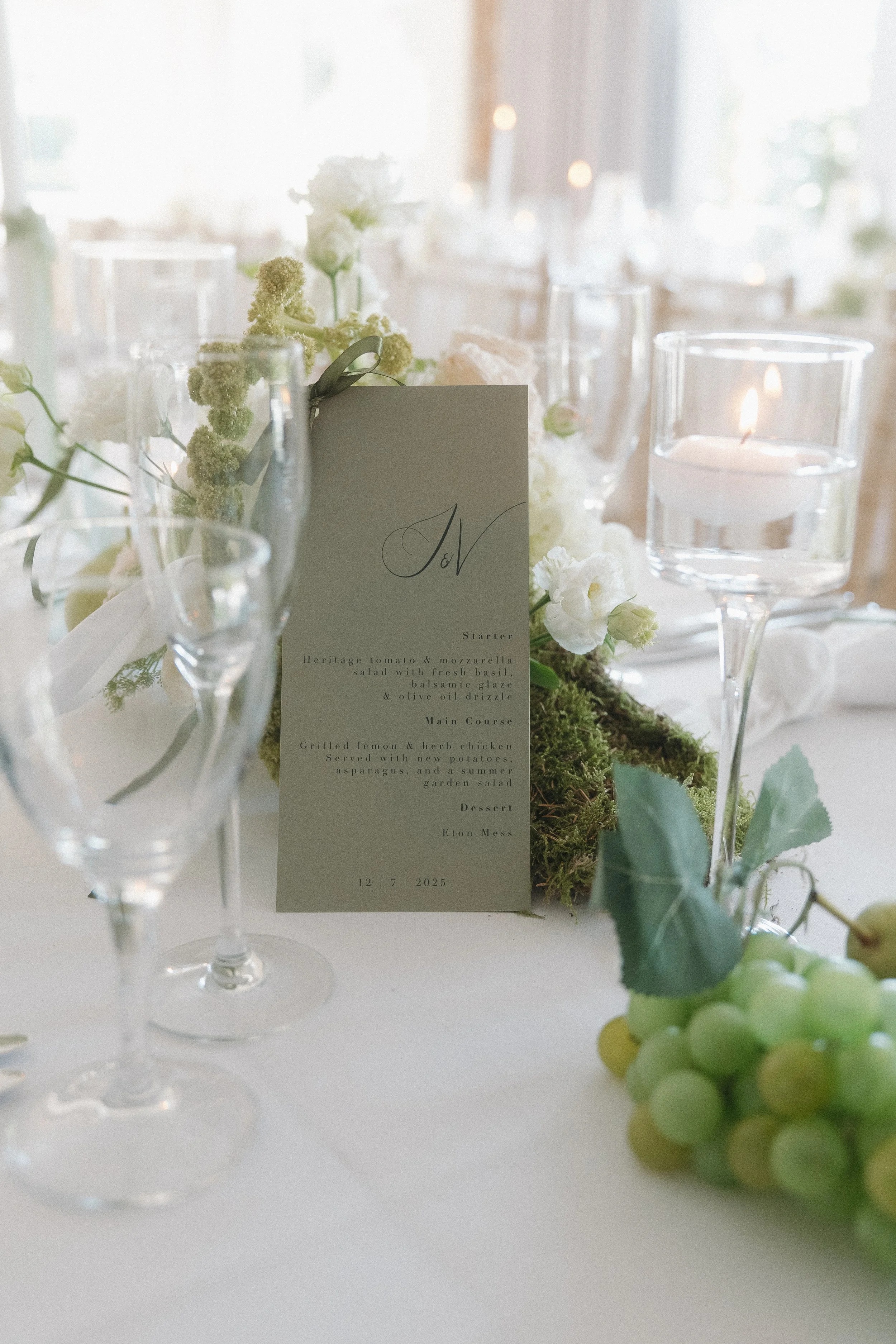 A dressed wedding table with white linens, glass tall candle holders and candles. Grapes have been used to dress the table. The centrepiece is a moss green menu stood up with callligraphy monogram details
