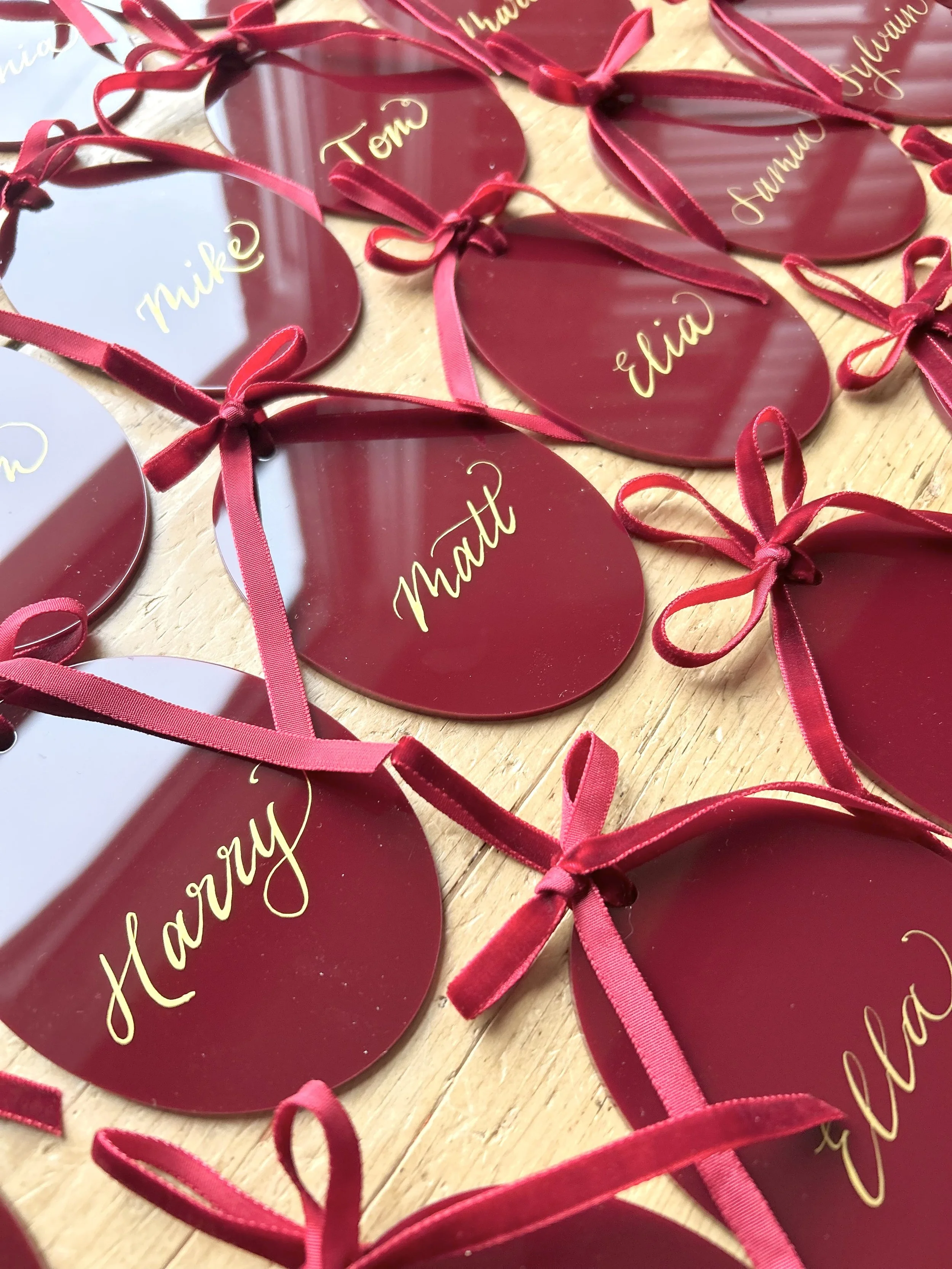 burgundy maroon acrylic place cards (oval shaped) with gold metallic lettering and tied with a velvet ribbon bow and loop to hang. a selection of acrylics placed on a wooden table