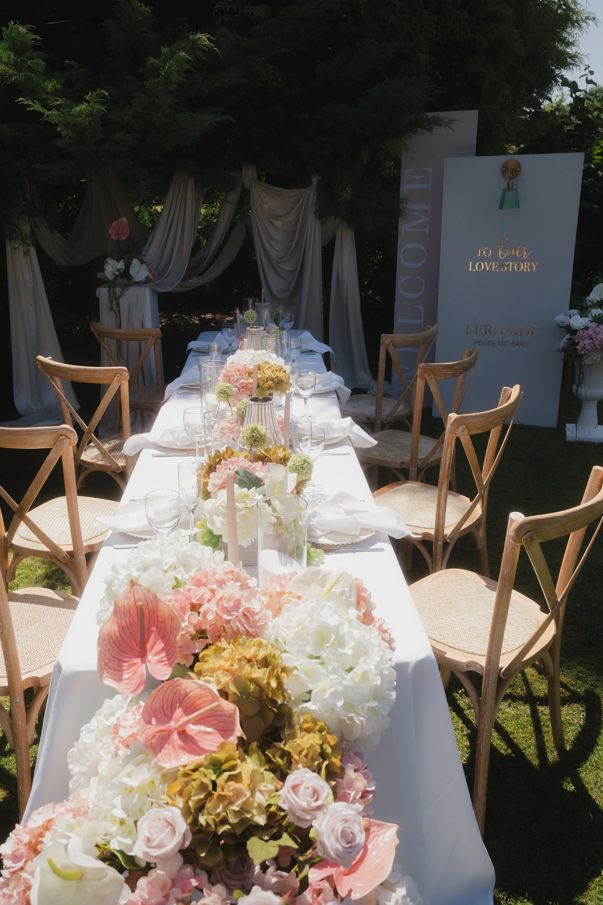 Outside wedding table set up with white and pink flowers cascading down. Large wooded signage under the trees
