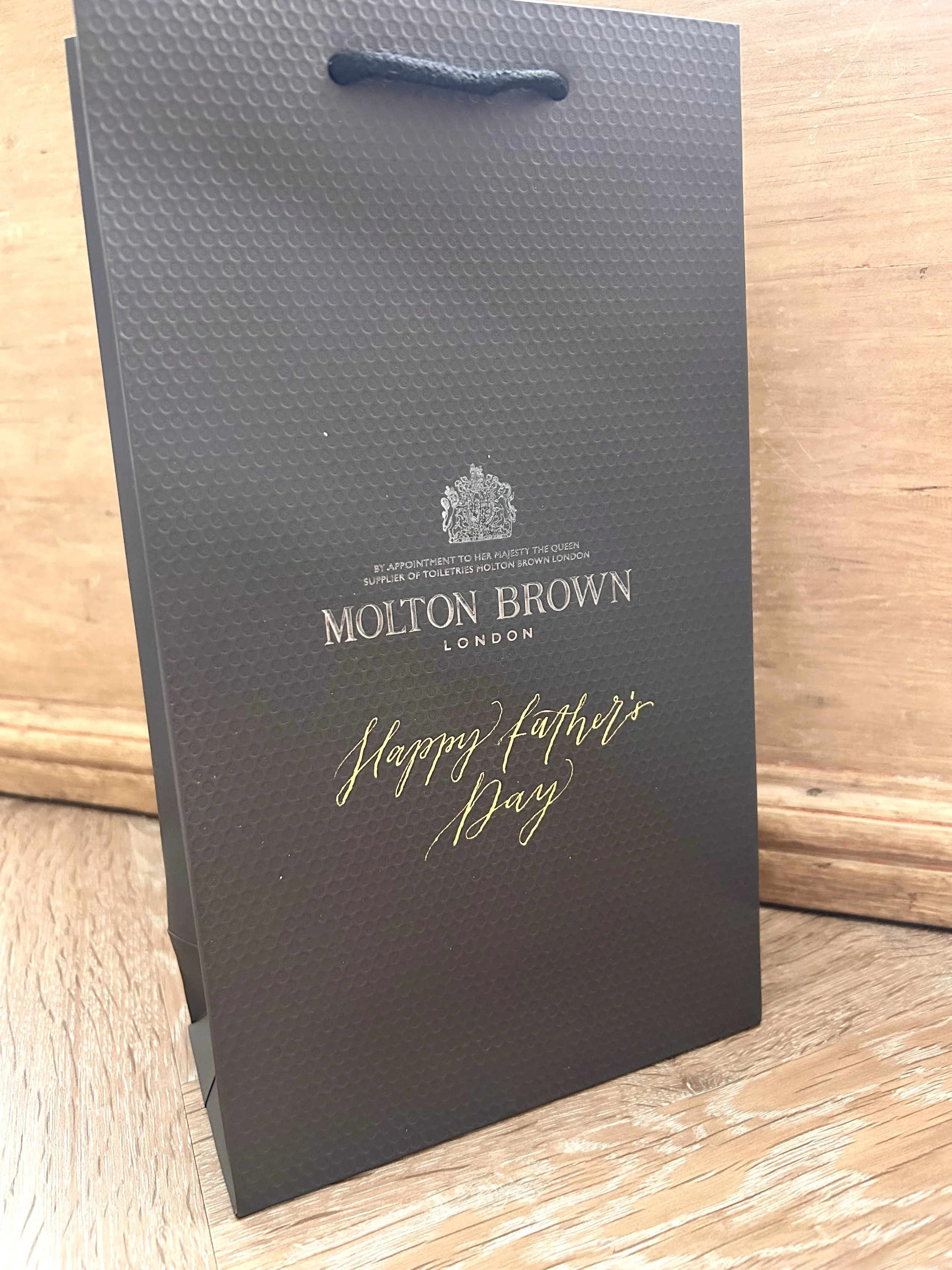 A molten brown gift bag (brown, textured) with calligraphy writing in gold below the logo- Happy father's day