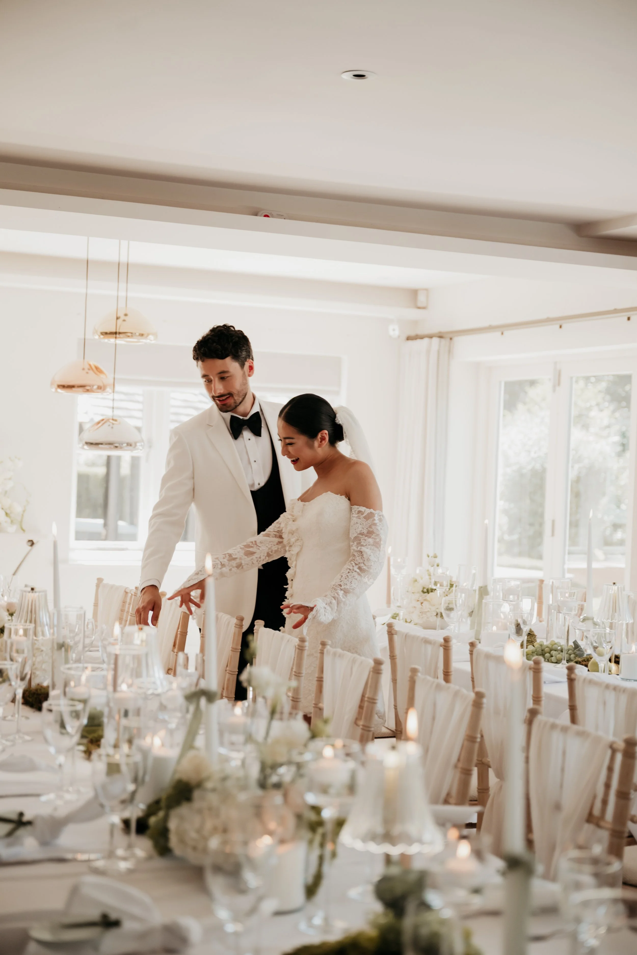 A bride and groom celebrating at their wedding reception, both smiling and reaching for a candle on the table, surrounded by elegant wine glasses, floral arrangements, and candles in a bright, decorated room.