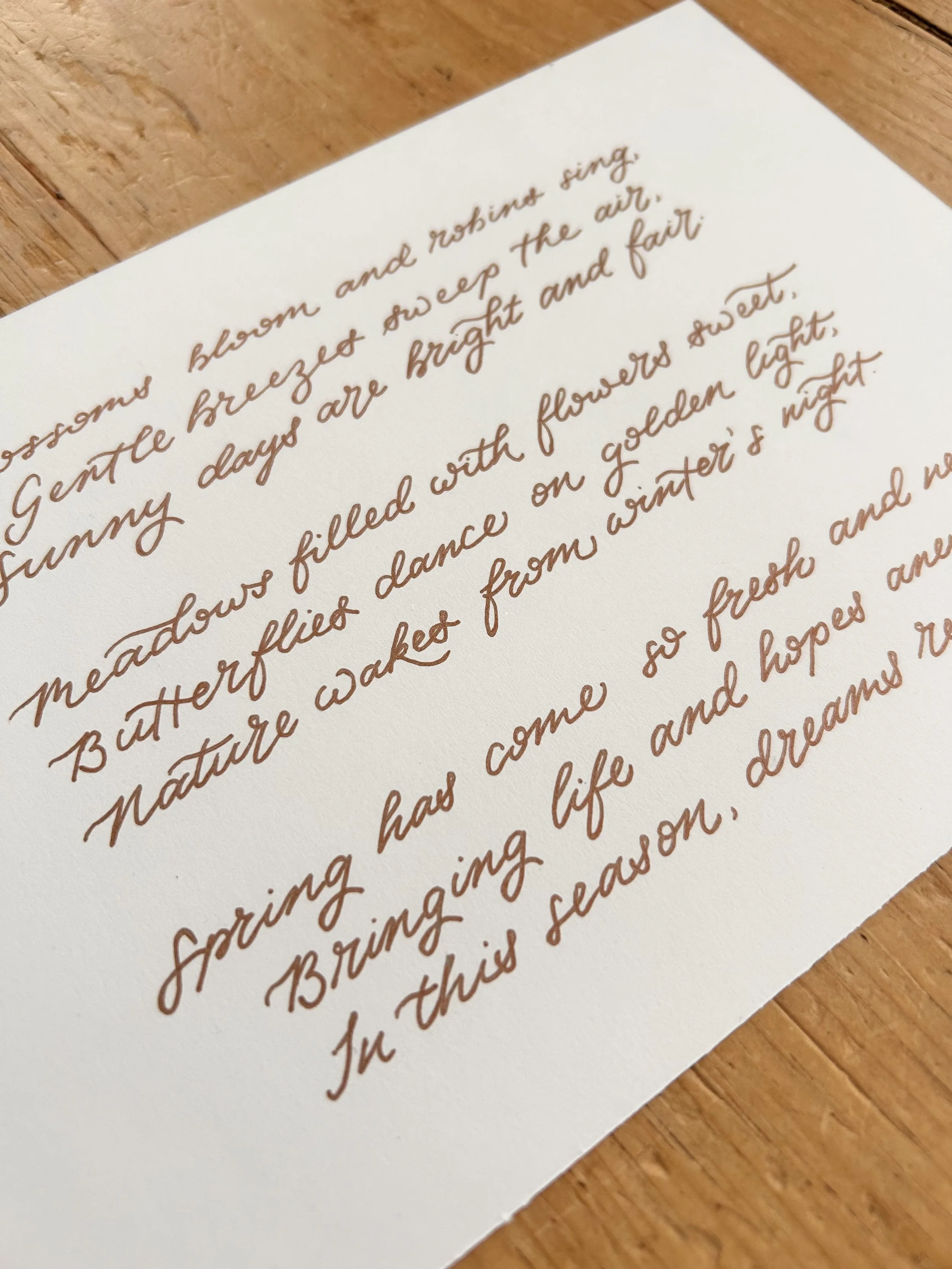 A white card with a flourished style handwriting in gold pen. The writing is a spring poem and is resting on a wooden table