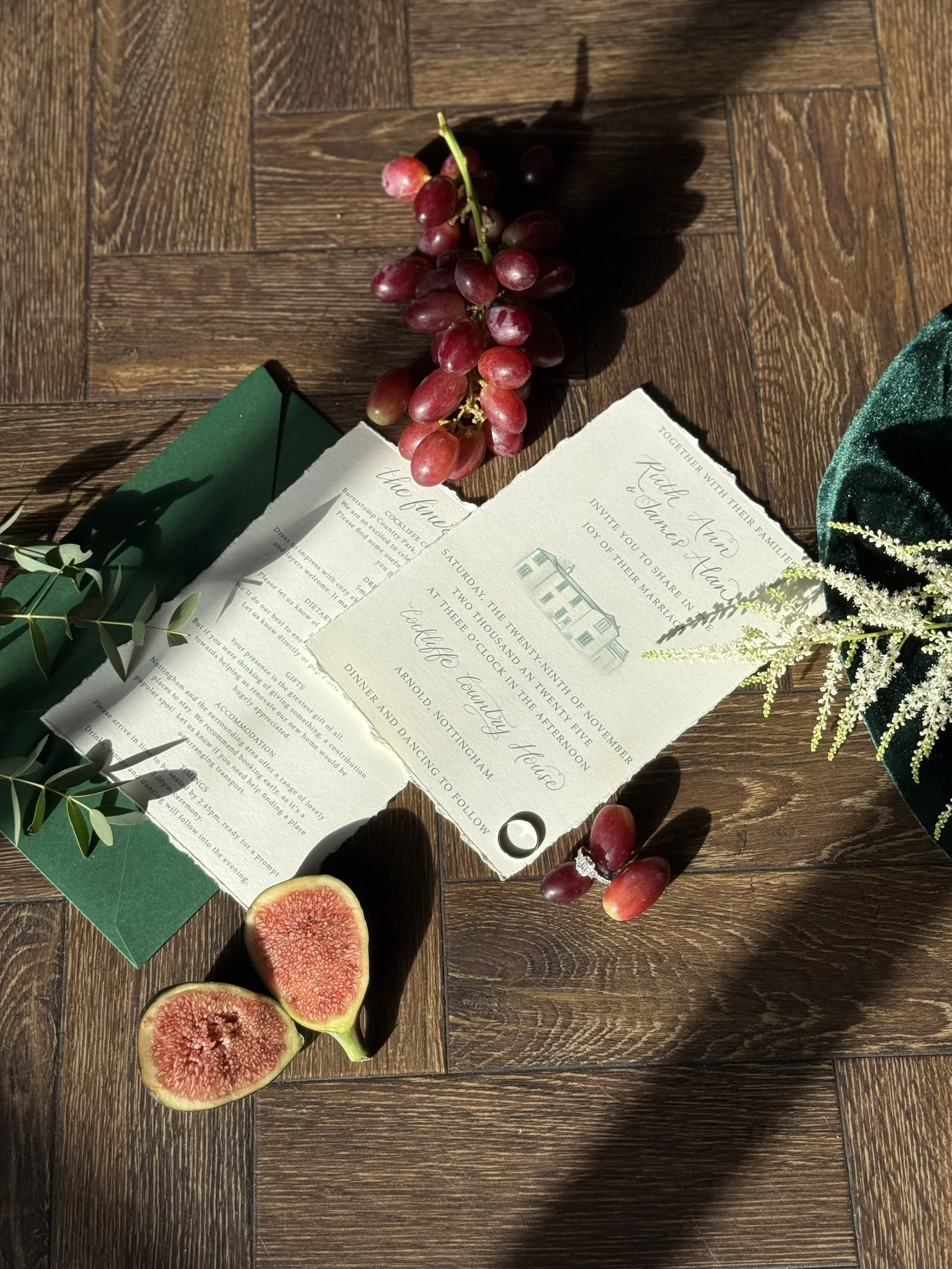 a flatlay image of a cream wedding invitation suite with green lettering and calligraphy details. it is accessorised with green envelope, foliage, grapes and a wedding ring. sunlight is shining onto the invitation