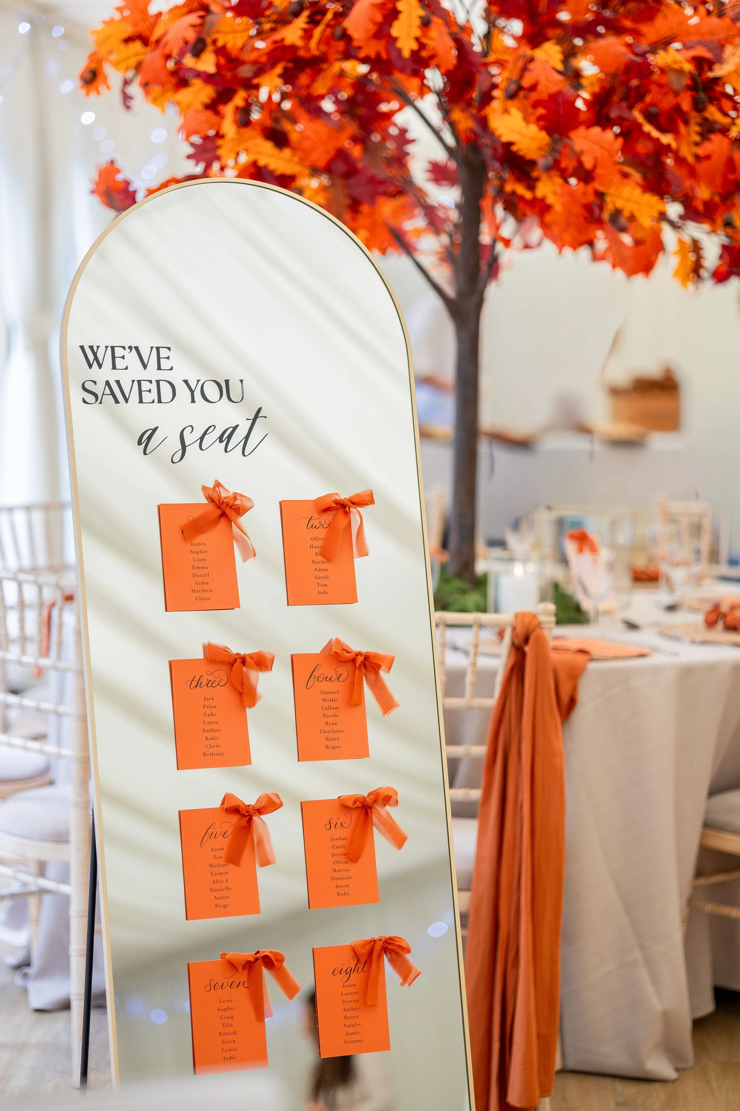 an autumn wedding set up with a table and organse leaved tree. In the foreground is a tall mirror with the wording 'we've saved you a seat'. The mirror has 8 burnt orange cards with calligraphy and table numbers accompanied by burnt orange ribbon