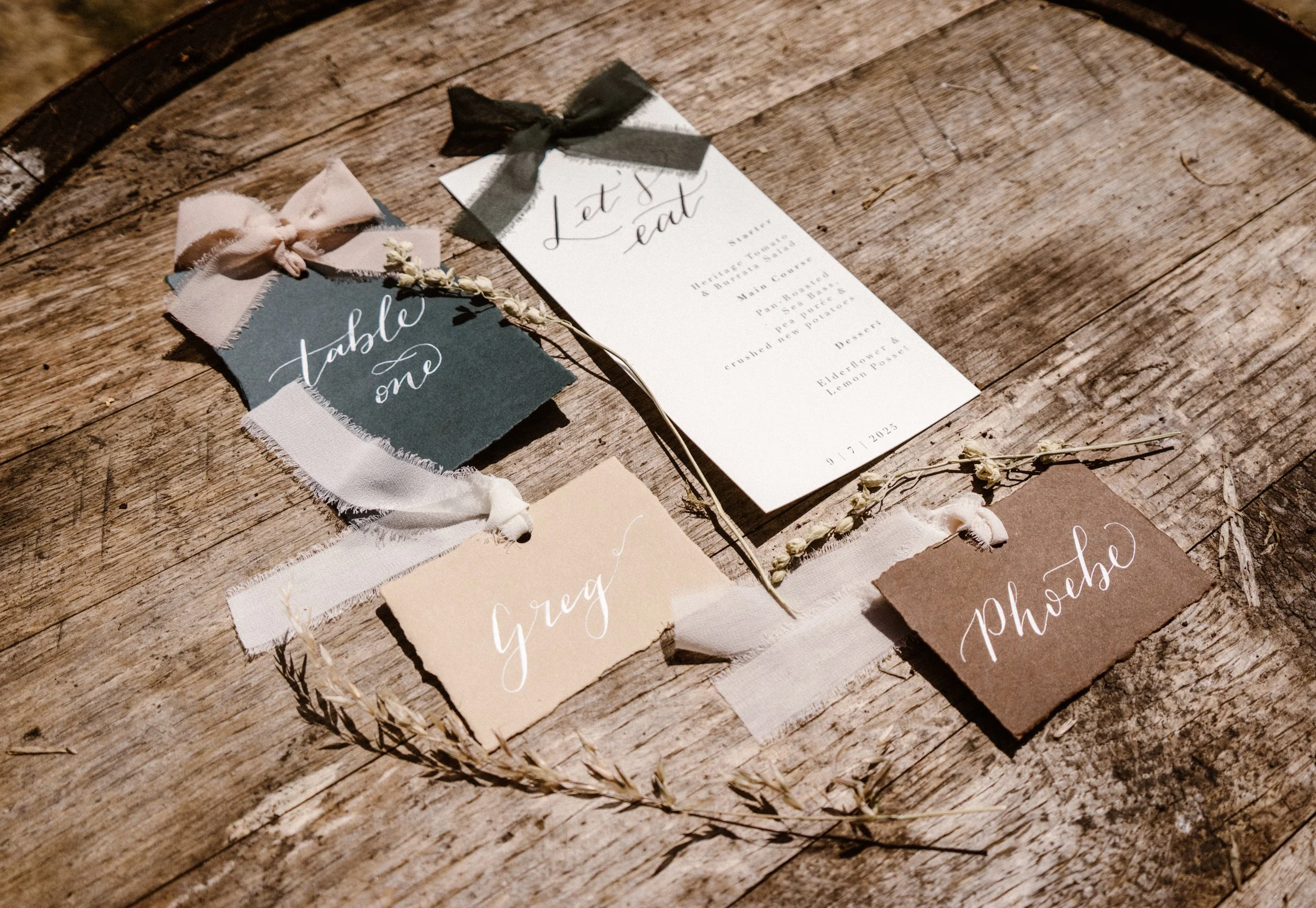 A rustic wedding stationery setup on a wooden surface, including a menu card, a table number tag with pink ribbon, a name tag labeled 'Greg' with white script, and a photo card labeled 'Photos.' The items are decorated with small dried flowers and fabric ribbons.