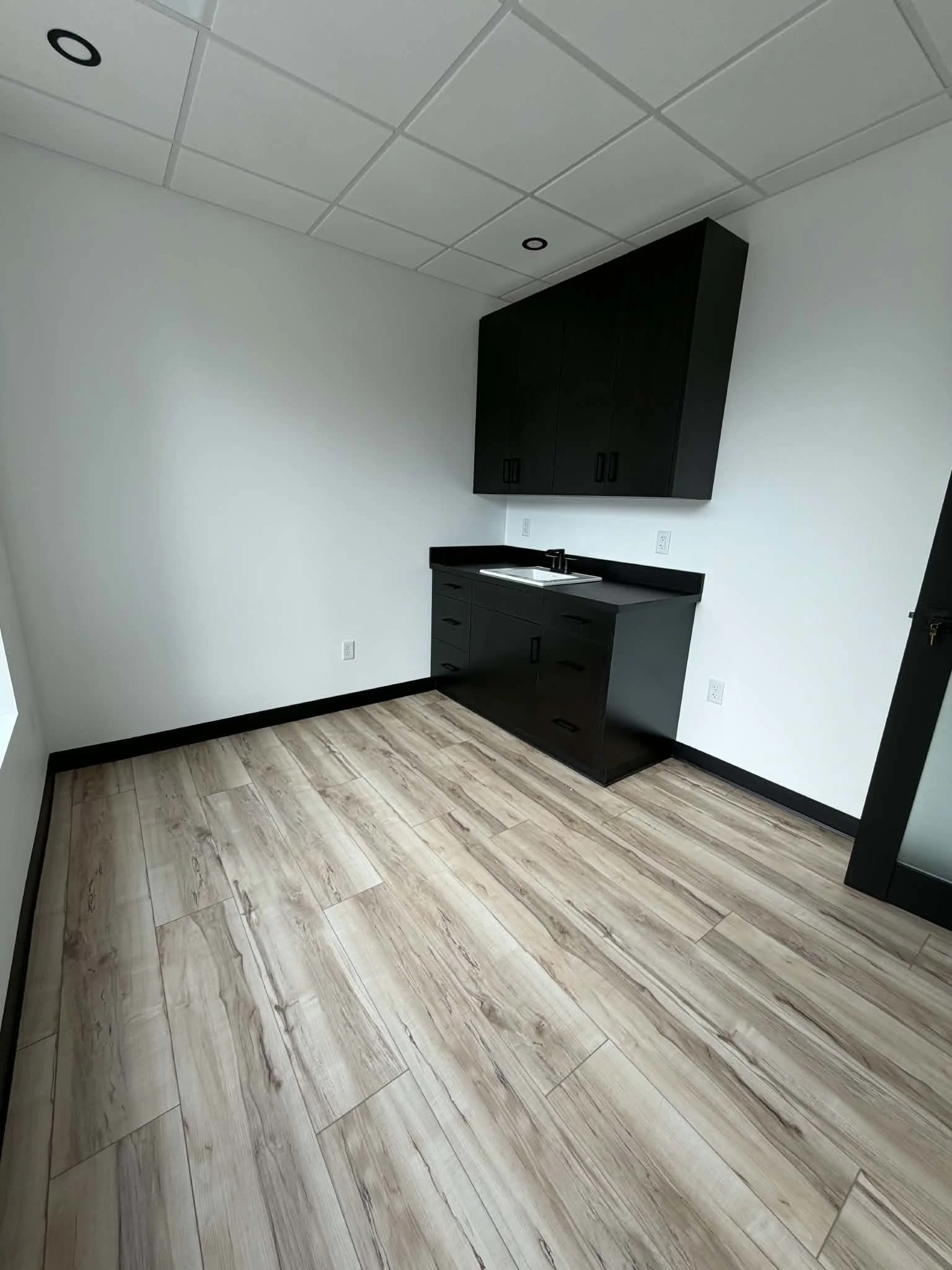 Empty room with light-colored wooden flooring, white walls, black cabinetry with a sink, and a white ceiling with square tiles and black circular lights.